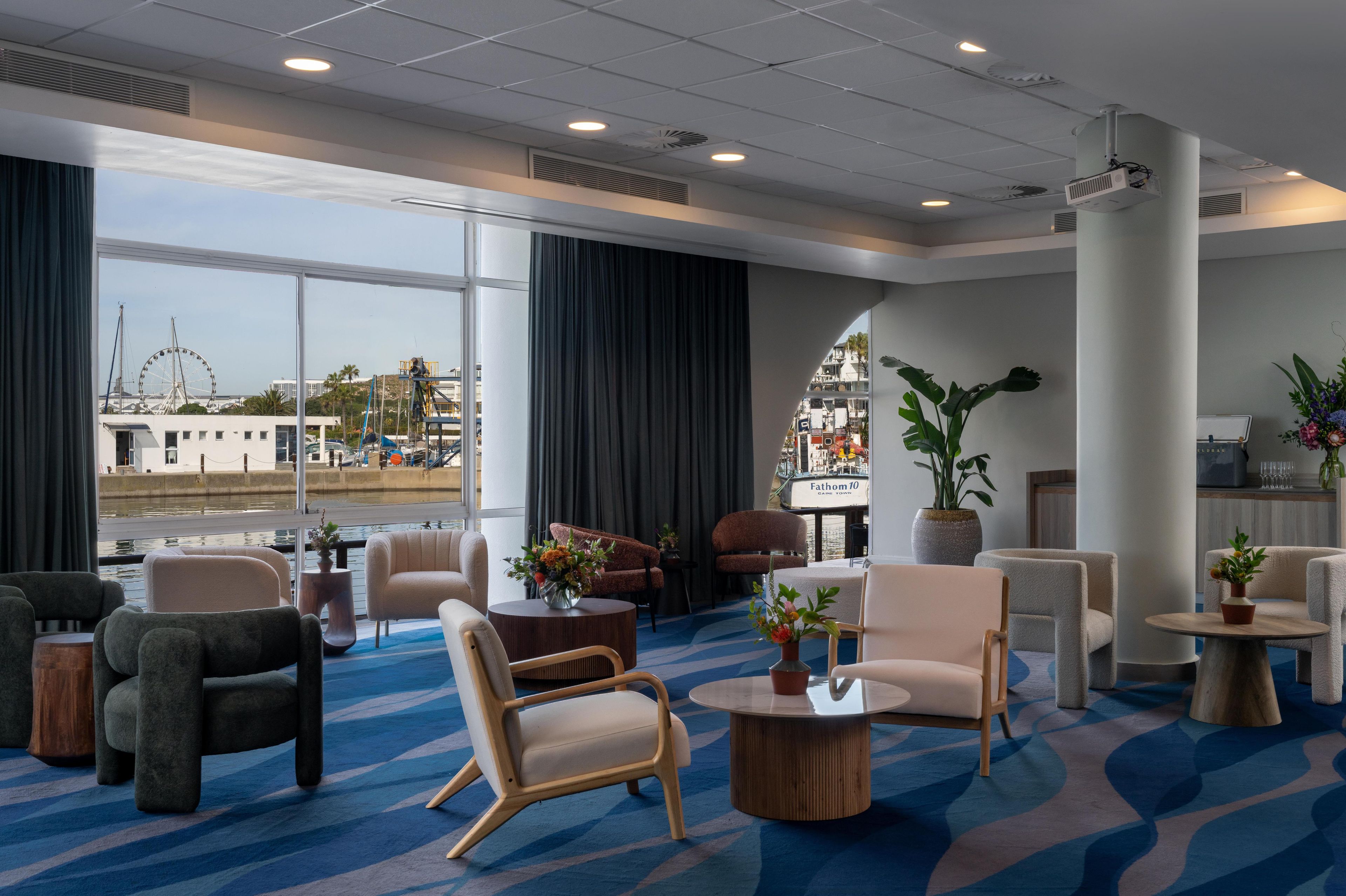 A hotel lobby with a large window offering a view of a waterfront and a Ferris wheel, furnished with modern chairs and tables, and a large pillar with a sign.