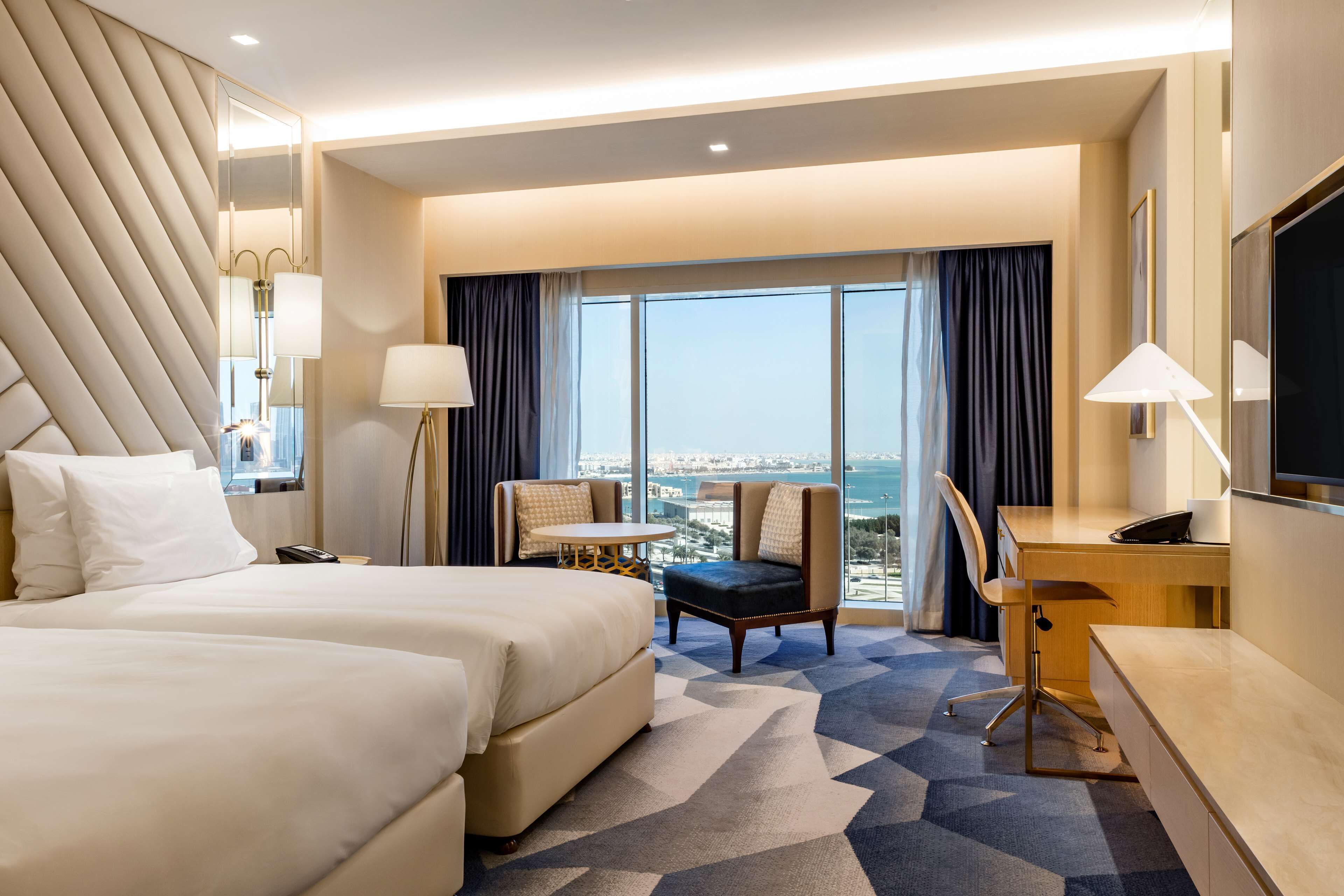 A hotel room with two beds, a desk, a chair, and a large window with a city view.