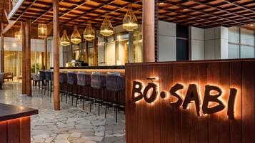 Interior de un restaurante con techo de madera, lámparas colgantes y una zona de bar con sillas y mesas.