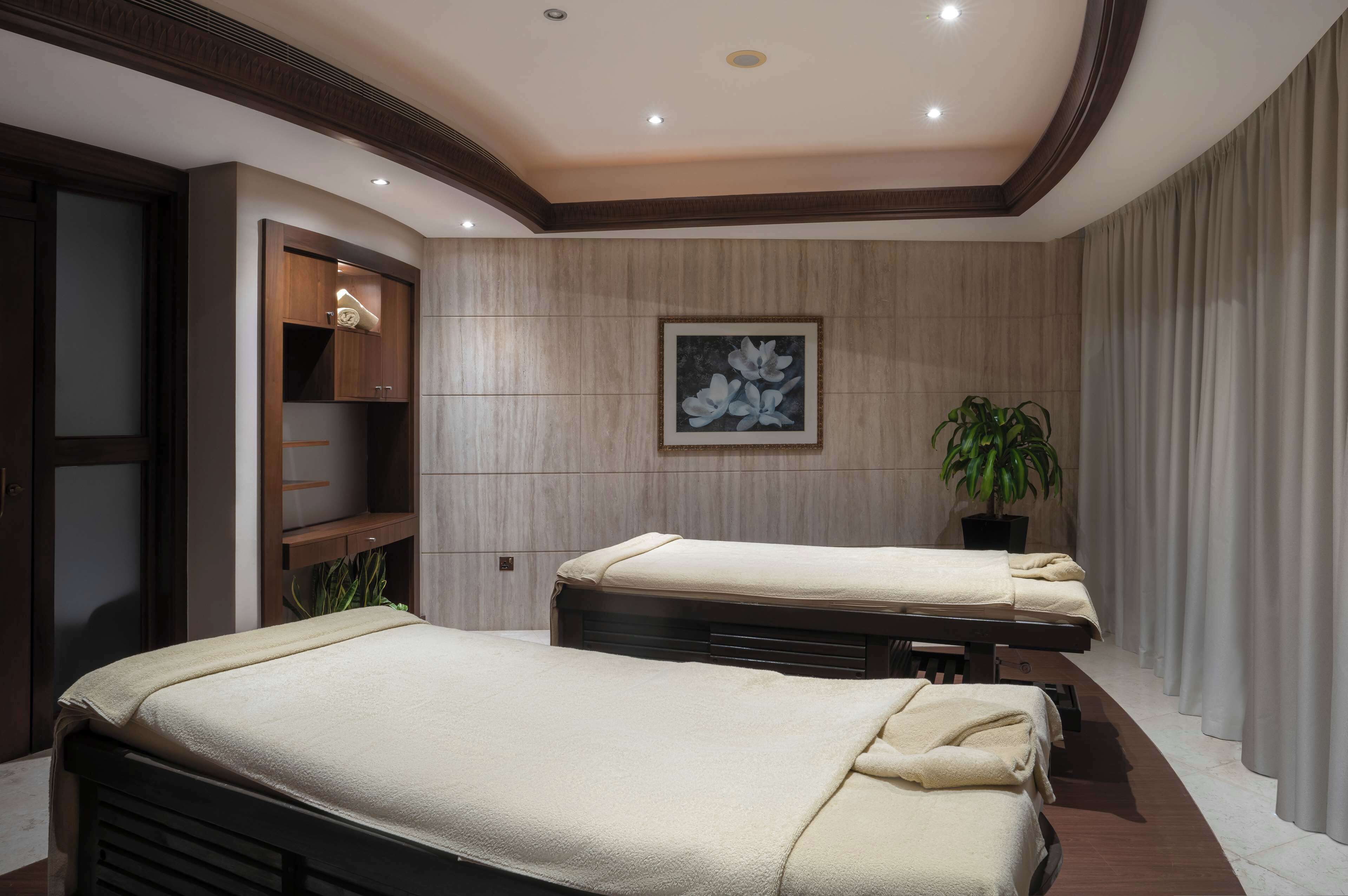 A spa room with two massage tables, a wooden cabinet, a framed floral artwork, and a potted plant.