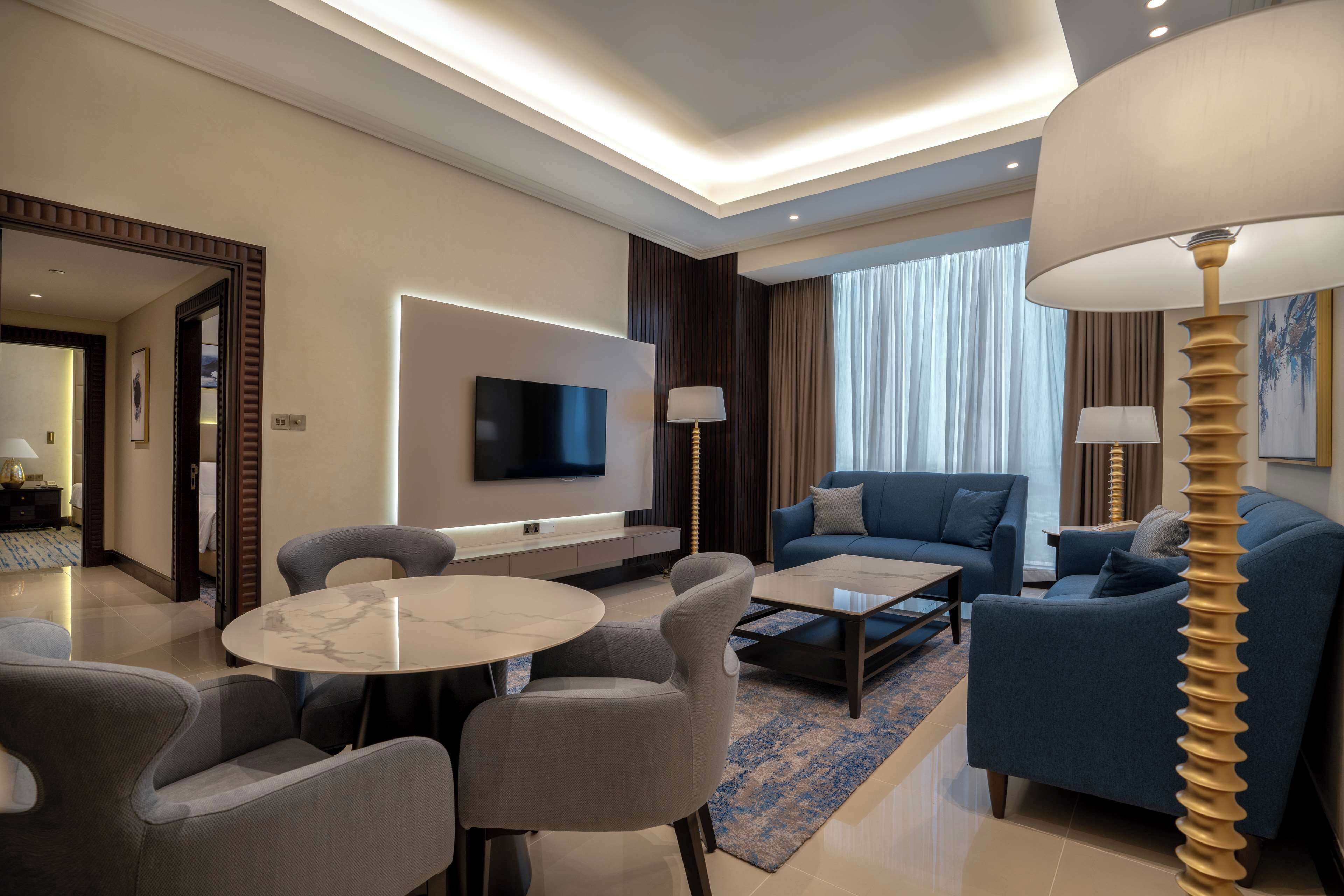 A modern hotel room with a flat-screen TV, a sofa, a dining table with chairs, and a lamp.