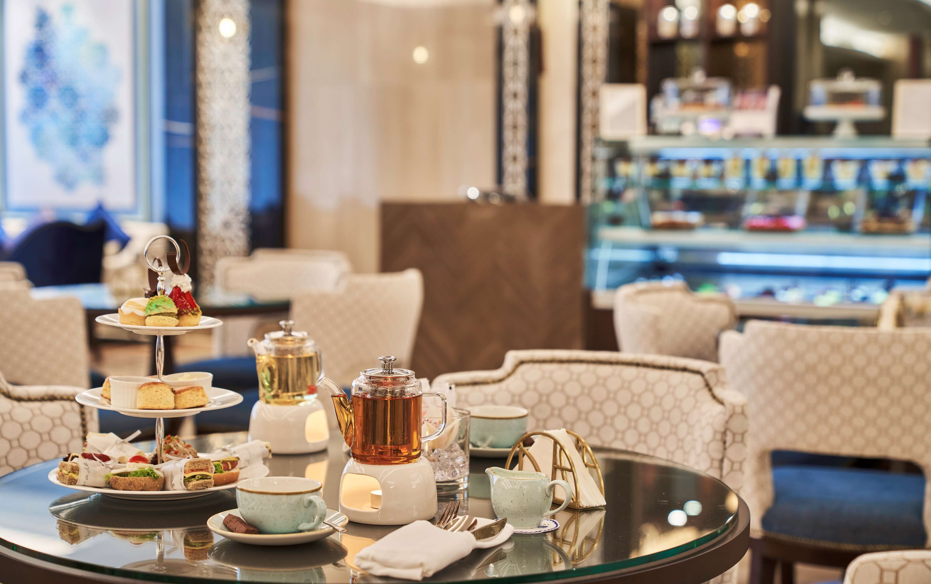 Une table ronde en verre, ornée d’un service à thé, de sandwichs et d’un présentoir à plusieurs niveaux pour les desserts, dans le hall d’un hôtel.