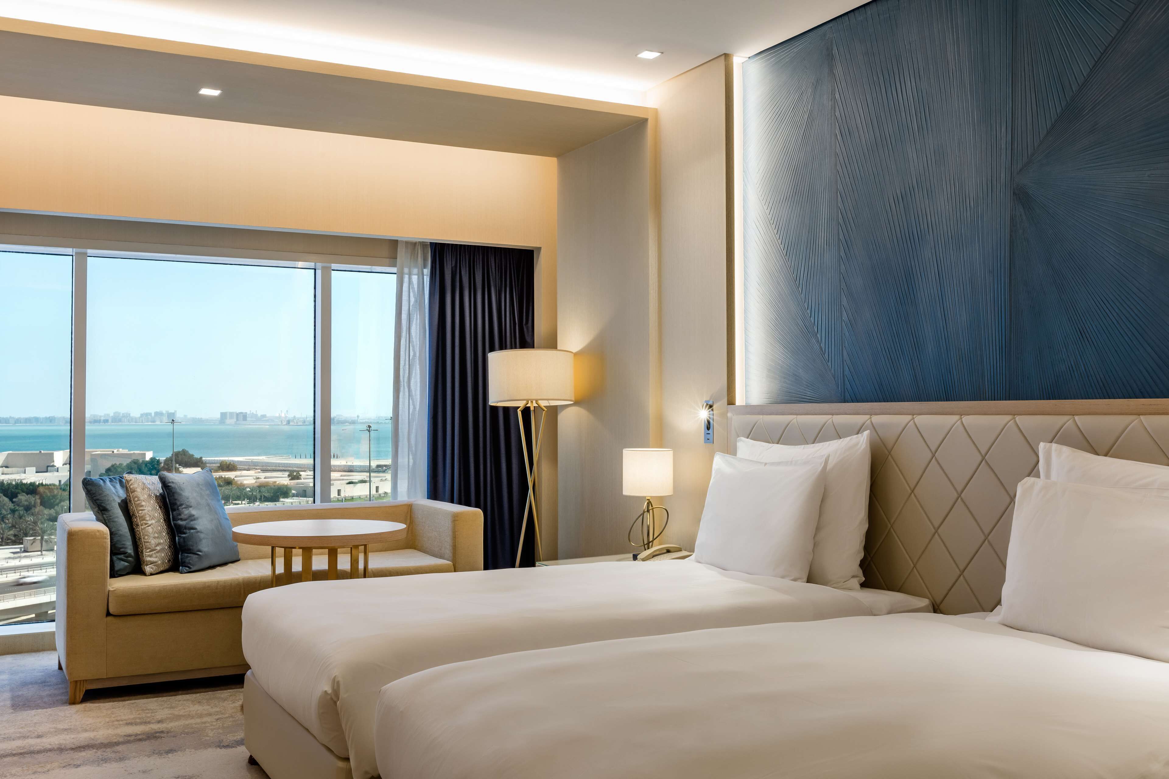 A hotel room with two beds, a sofa, a small round table, a lamp, and a large window offering a city view.