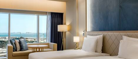 A hotel room with two beds, a sofa, a small round table, a lamp, and a large window offering a city view.