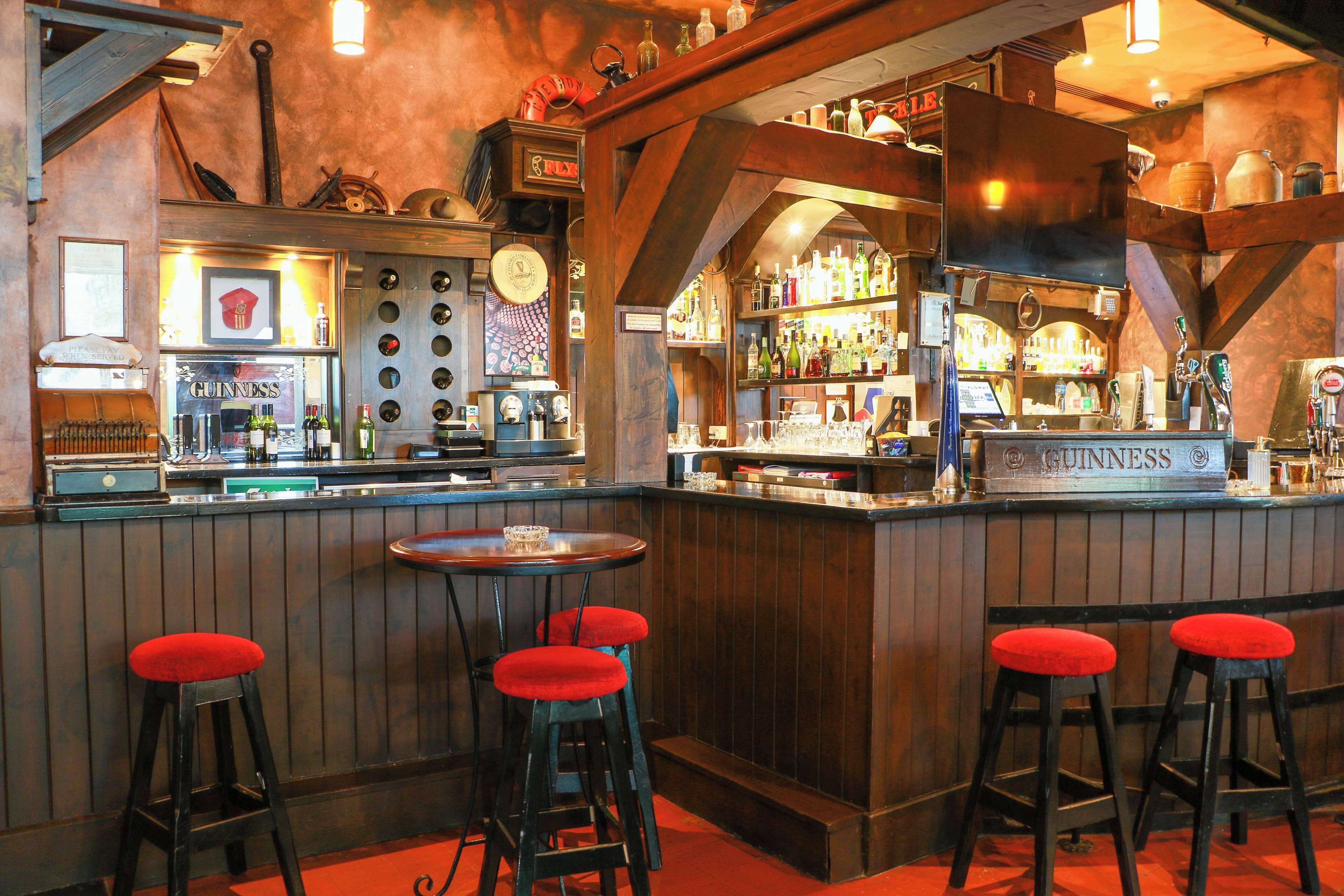 Una zona de bar con taburetes de madera, un letrero de Guinness y varias botellas y vasos en estantes.