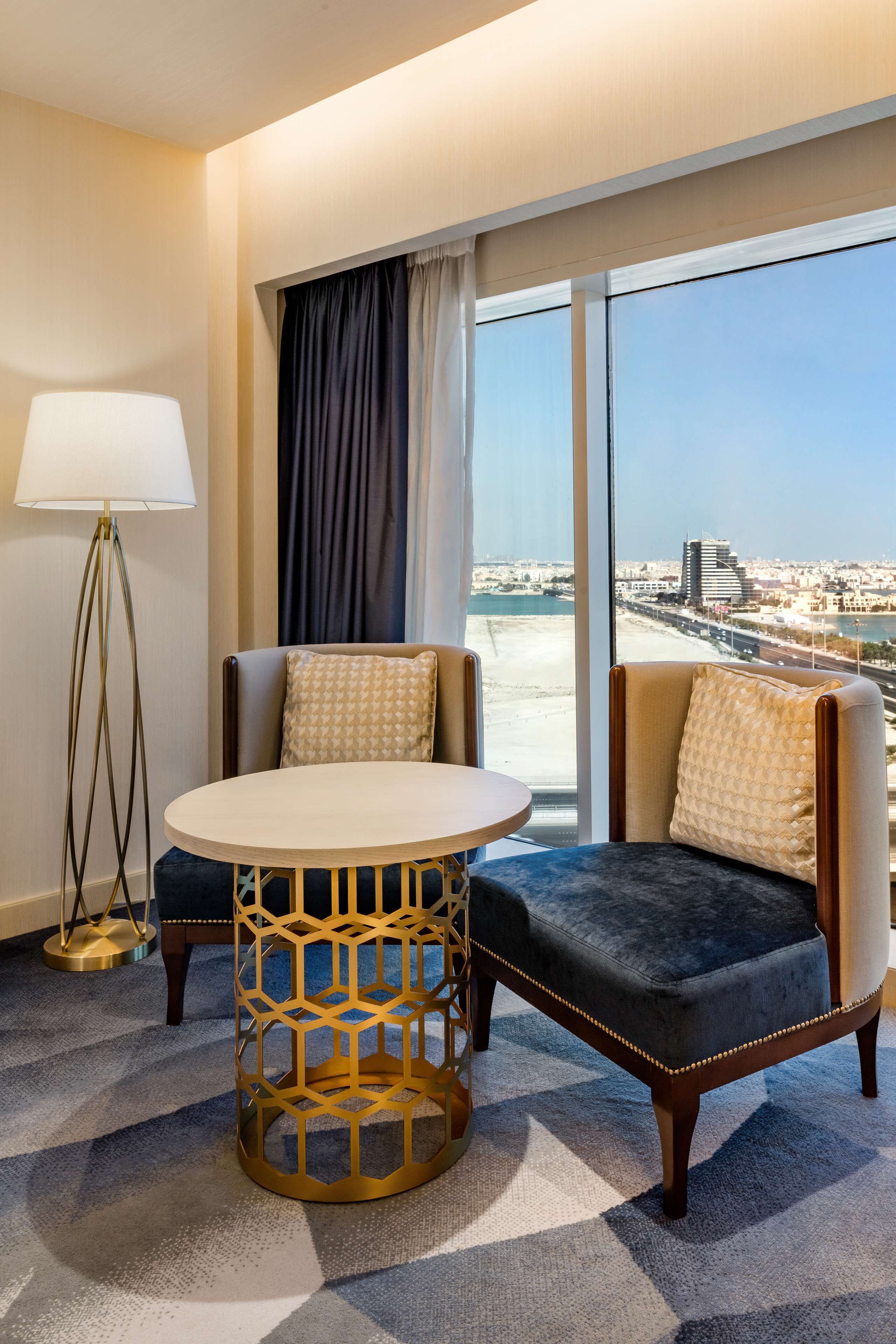 Hotelkamer met een groot raam, een ronde tafel, twee fauteuils, een vloerlamp en een kamerbreed tapijt met dessin.