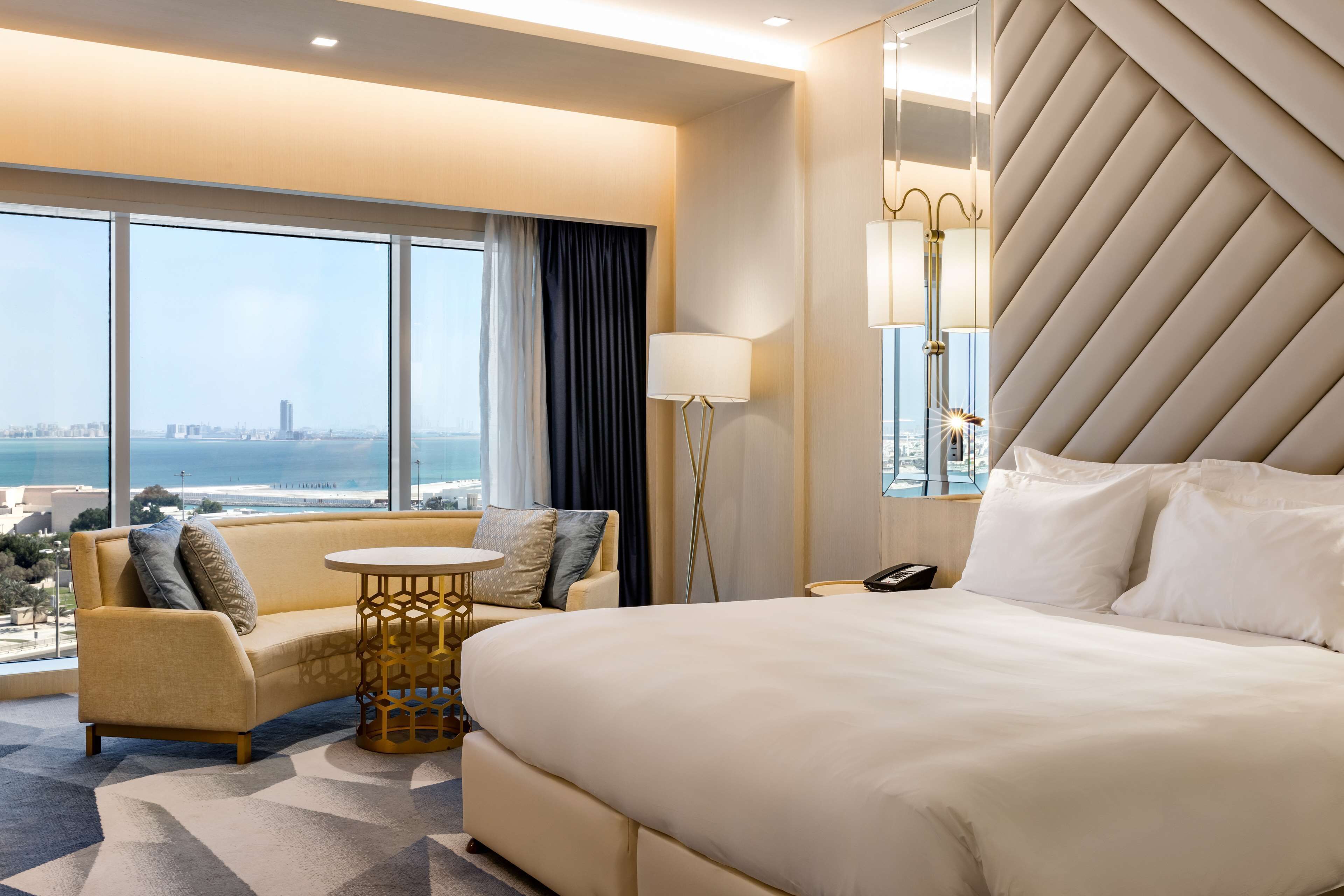 A hotel room with a large bed, a sofa, a small round table, and a large window offering a city view.