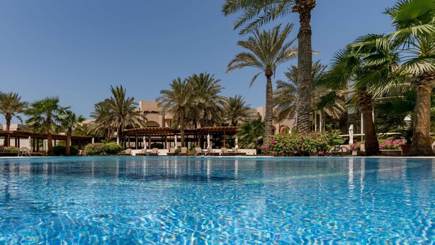 A clear blue pool surrounded by palm trees and lounge chairs, with a building in the background. - The Diplomat Radisson BLU Hotel, Residence & Spa (Manama)