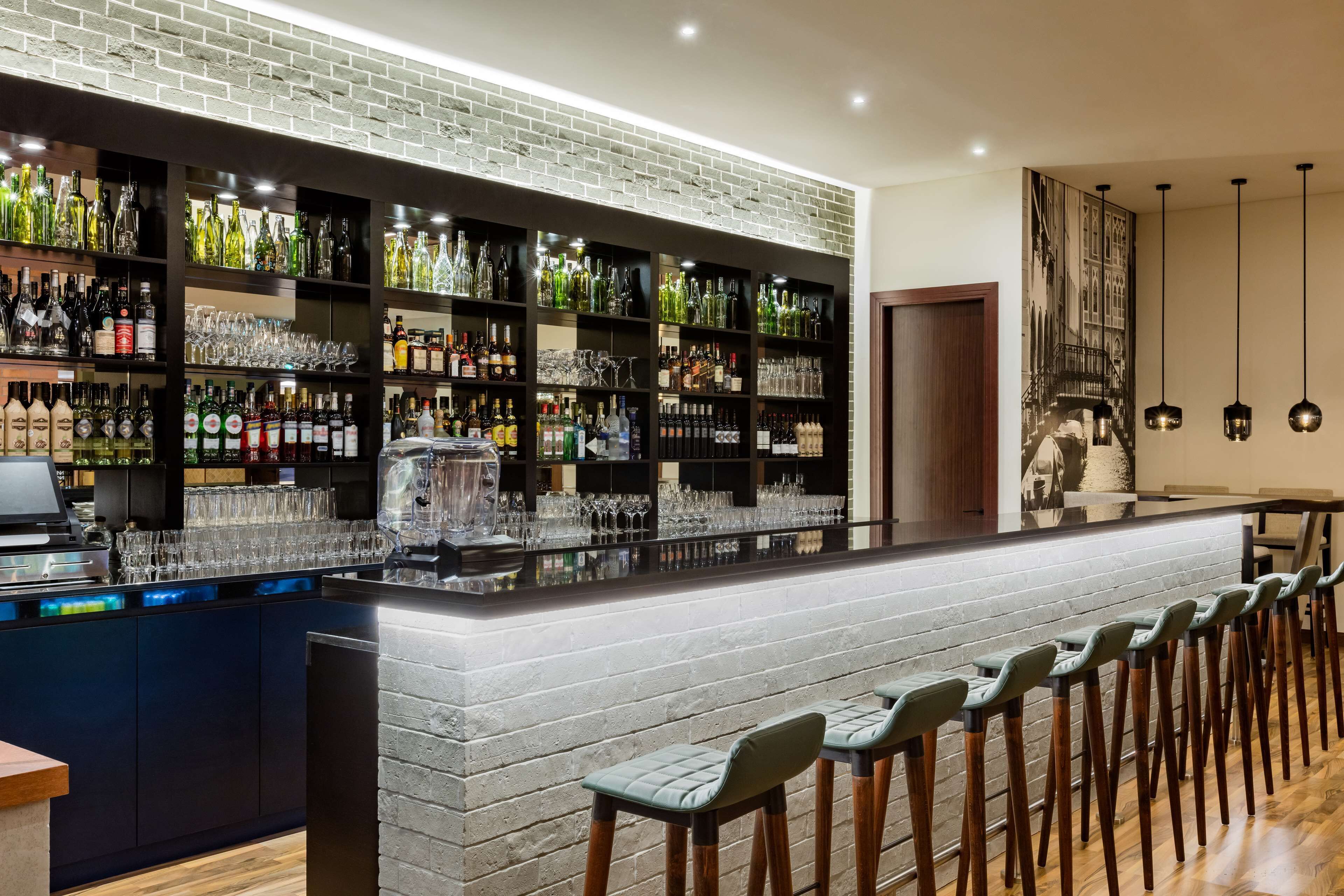 A bar area with a brick wall, shelves of bottles, glasses, and a long counter with bar stools.