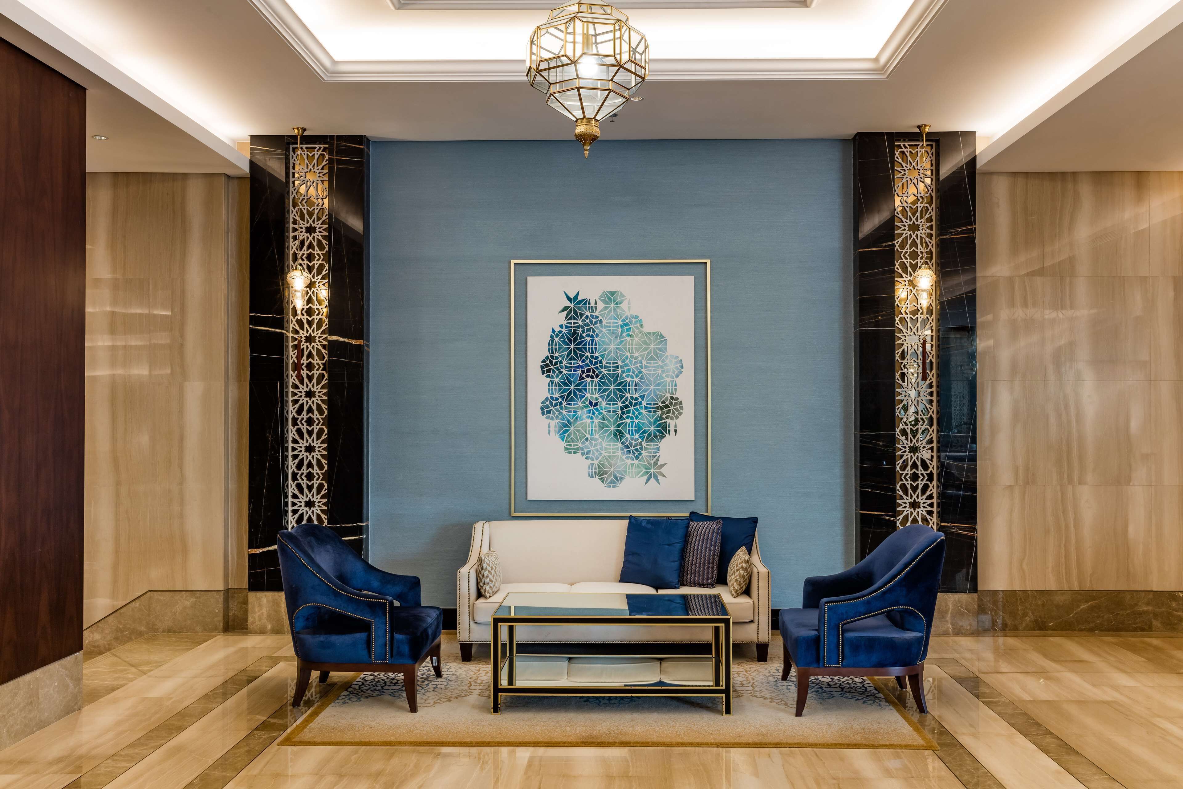 Eine Lobby mit blauer Wand, einem gerahmten abstrakten Kunstwerk, einem weißen Sofa, zwei blauen Sesseln und einem Glastisch.