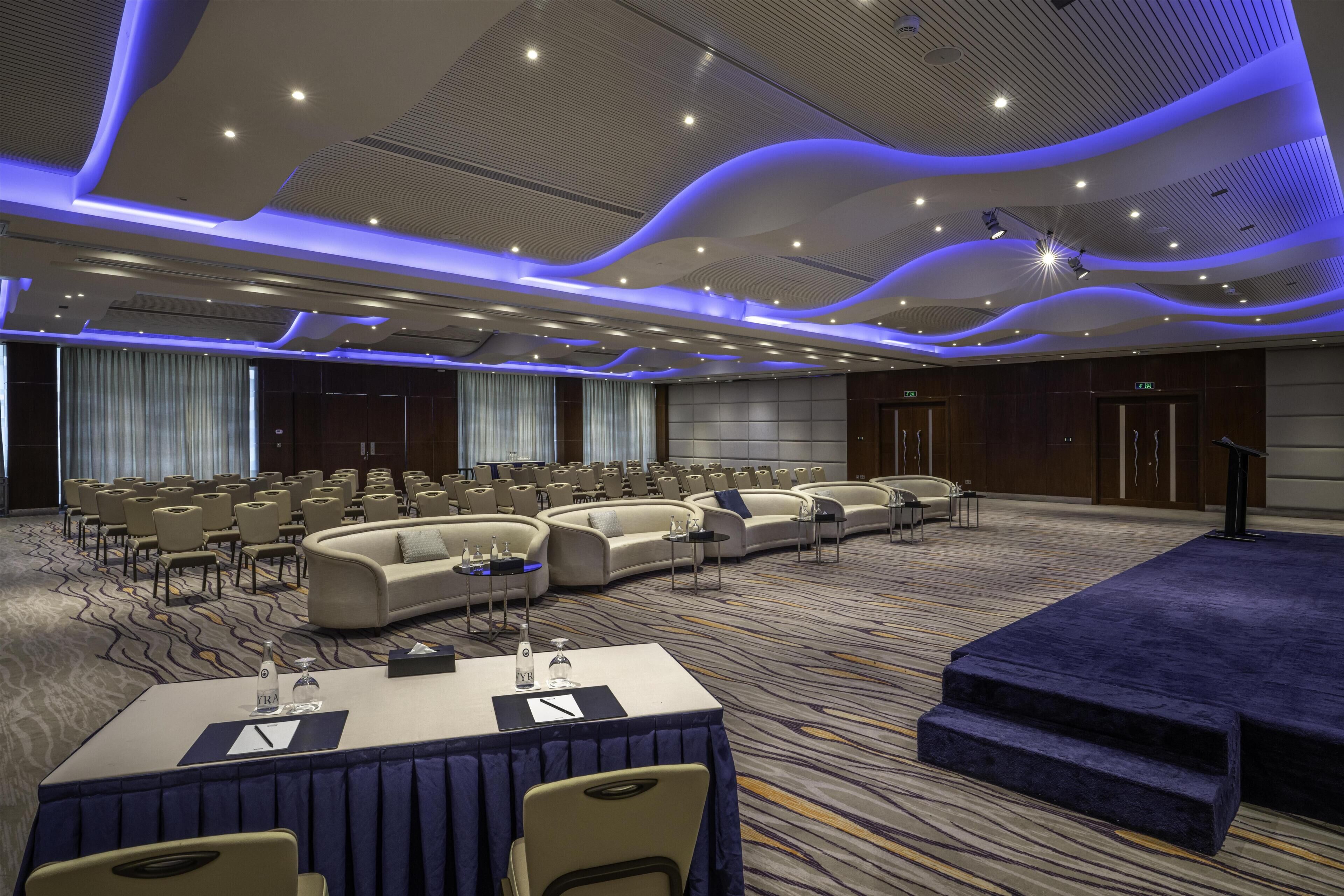 Un salón de conferencias con filas de sillas, un escenario y una zona de estar con sofás.