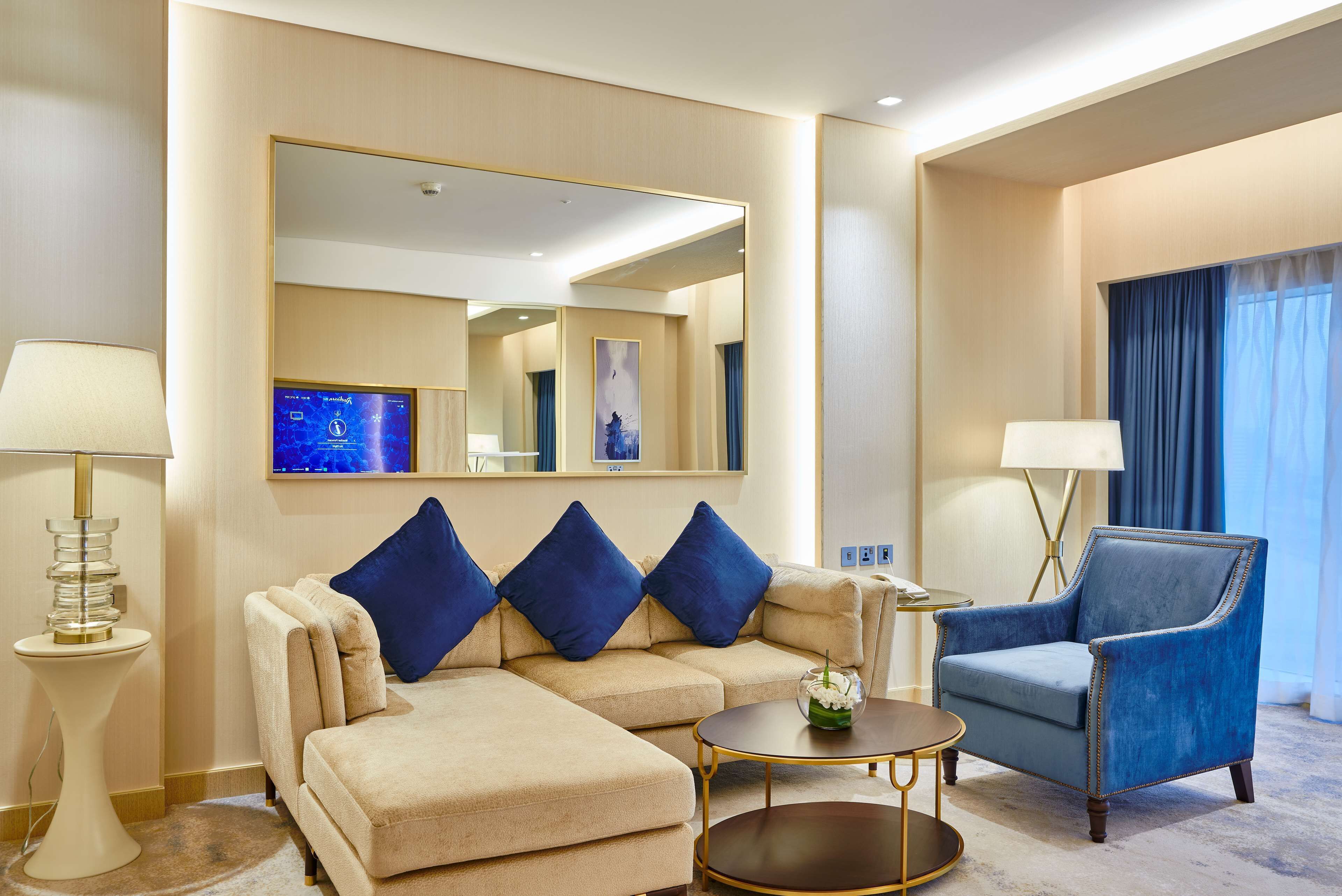 A hotel room with a beige sofa, a blue armchair, a round coffee table, a side table with a lamp, and a large mirror on the wall.
