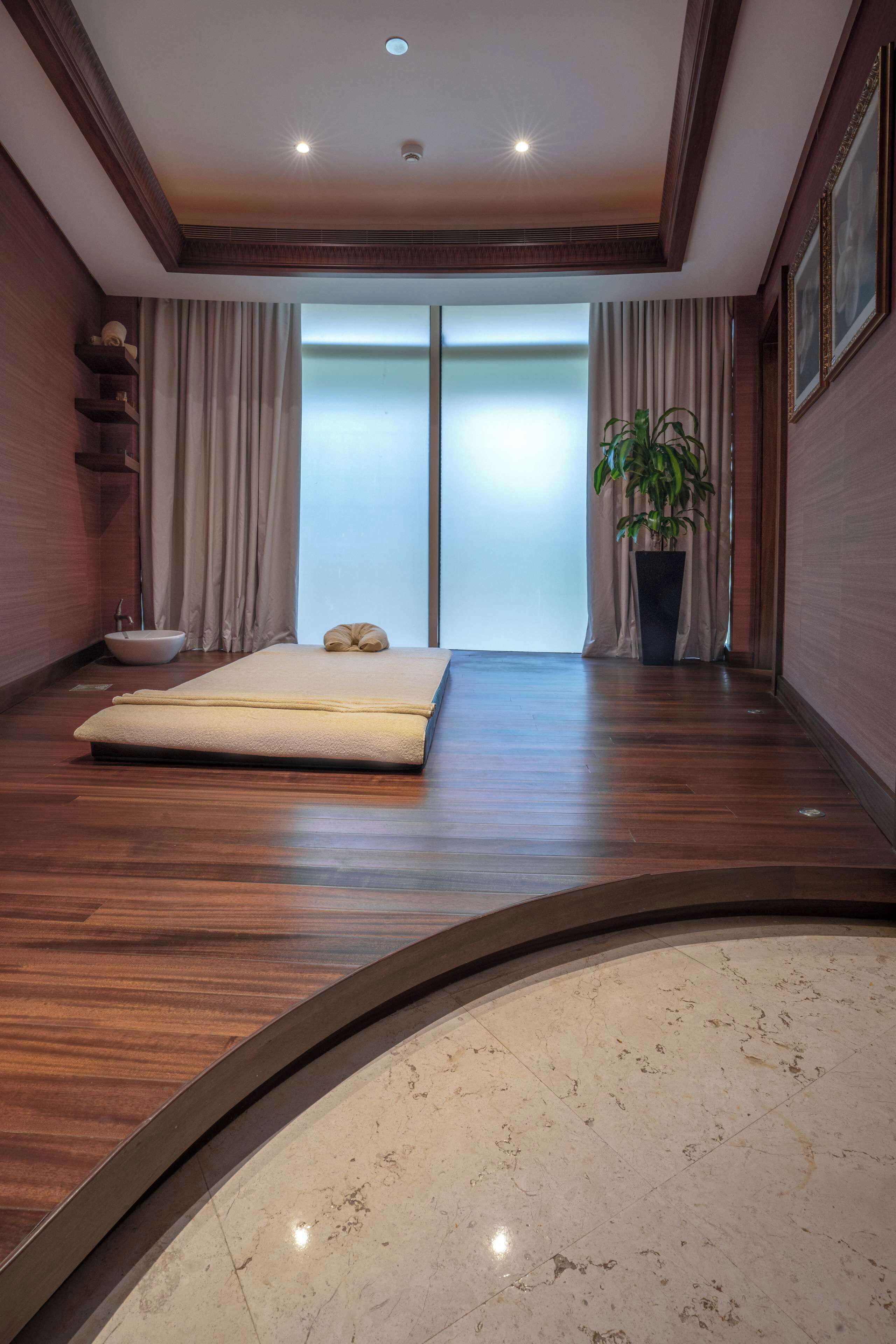 Una habitación con piso de madera, una cama con colchón blanco, una planta en maceta, un ventanal con cortinas y un lavamanos pequeño.