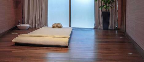 Una habitación con piso de madera, una cama con colchón blanco, una planta en maceta, un ventanal con cortinas y un lavamanos pequeño.