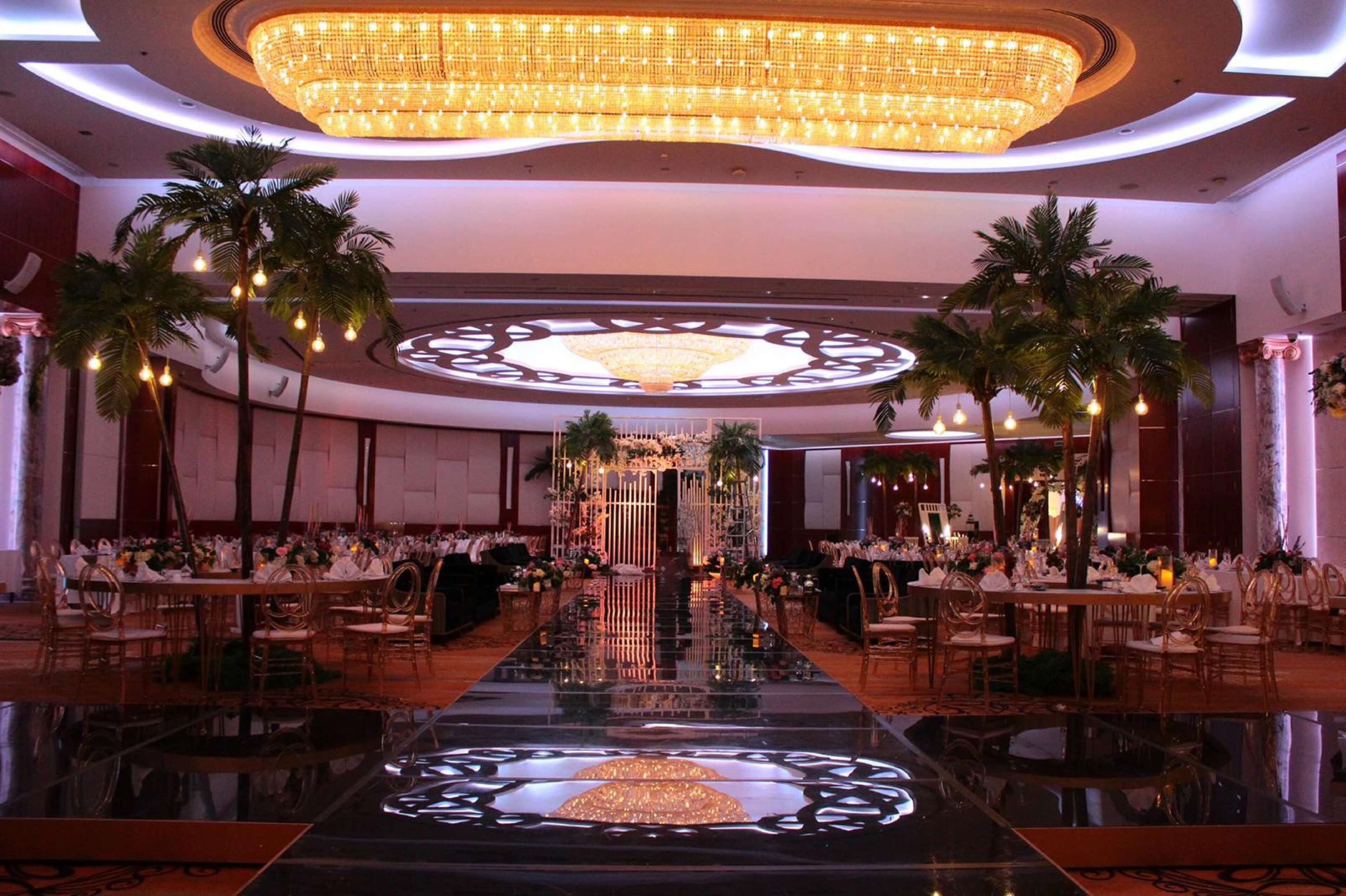 A banquet hall with a reflective floor, a central floral arrangement, and palm trees.