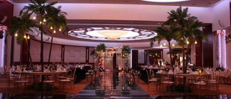 Un salón de banquetes con piso reflectante, un arreglo floral central y palmeras.