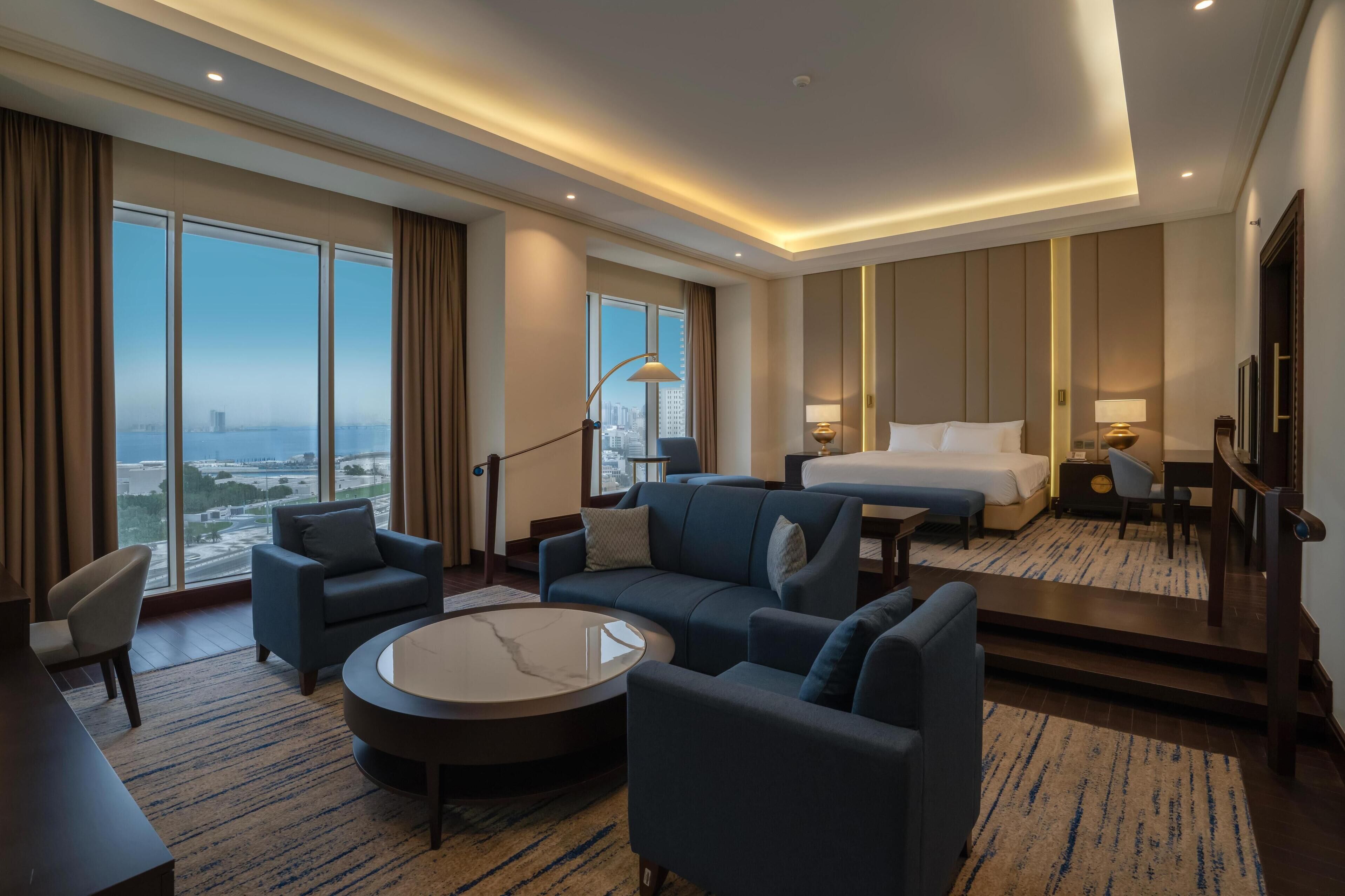 A modern hotel room with a large bed, two sofas, a coffee table, and a desk. The room has large windows with a city view.