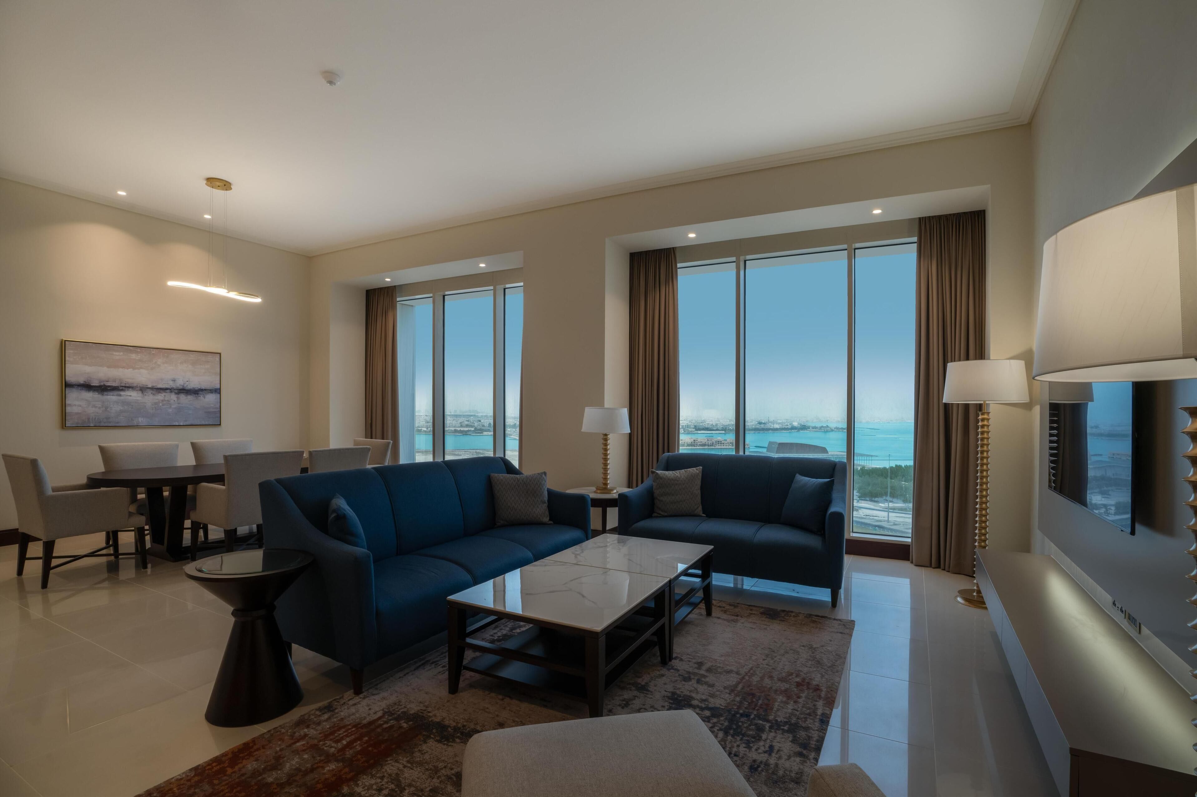 Una sala de estar moderna con dos sofás, una mesa de centro, una mesa de comedor y sillas. Amplias ventanas ofrecen vistas de la ciudad.
