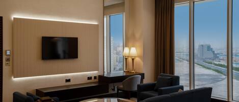A modern hotel room with a large TV mounted on a wall, a comfortable seating area with sofas and a coffee table, and a large window offering a city view.
