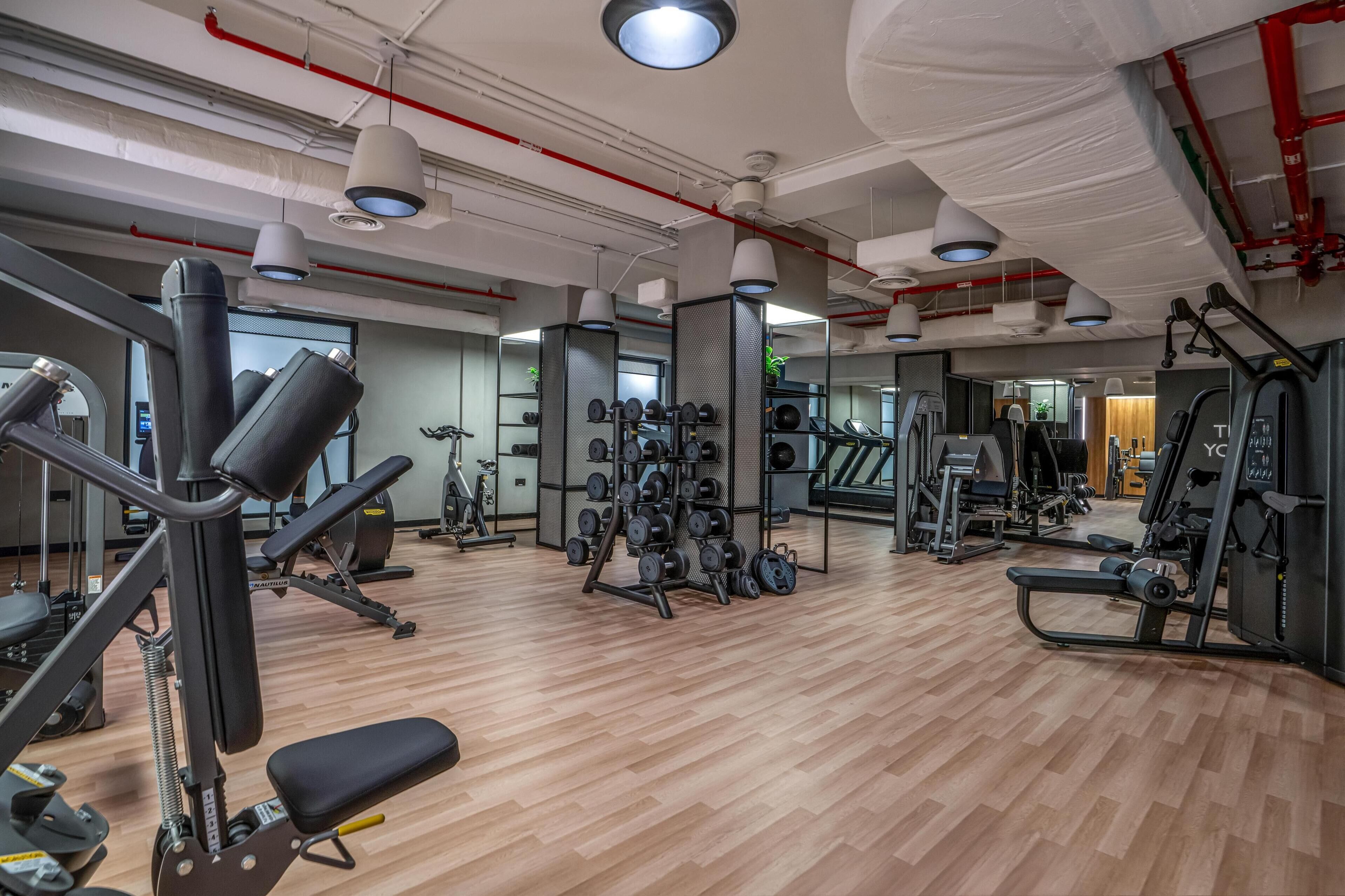 Une salle de sport moderne équipée de divers appareils d’exercice, dotée d’un plancher en bois et de grandes fenêtres.