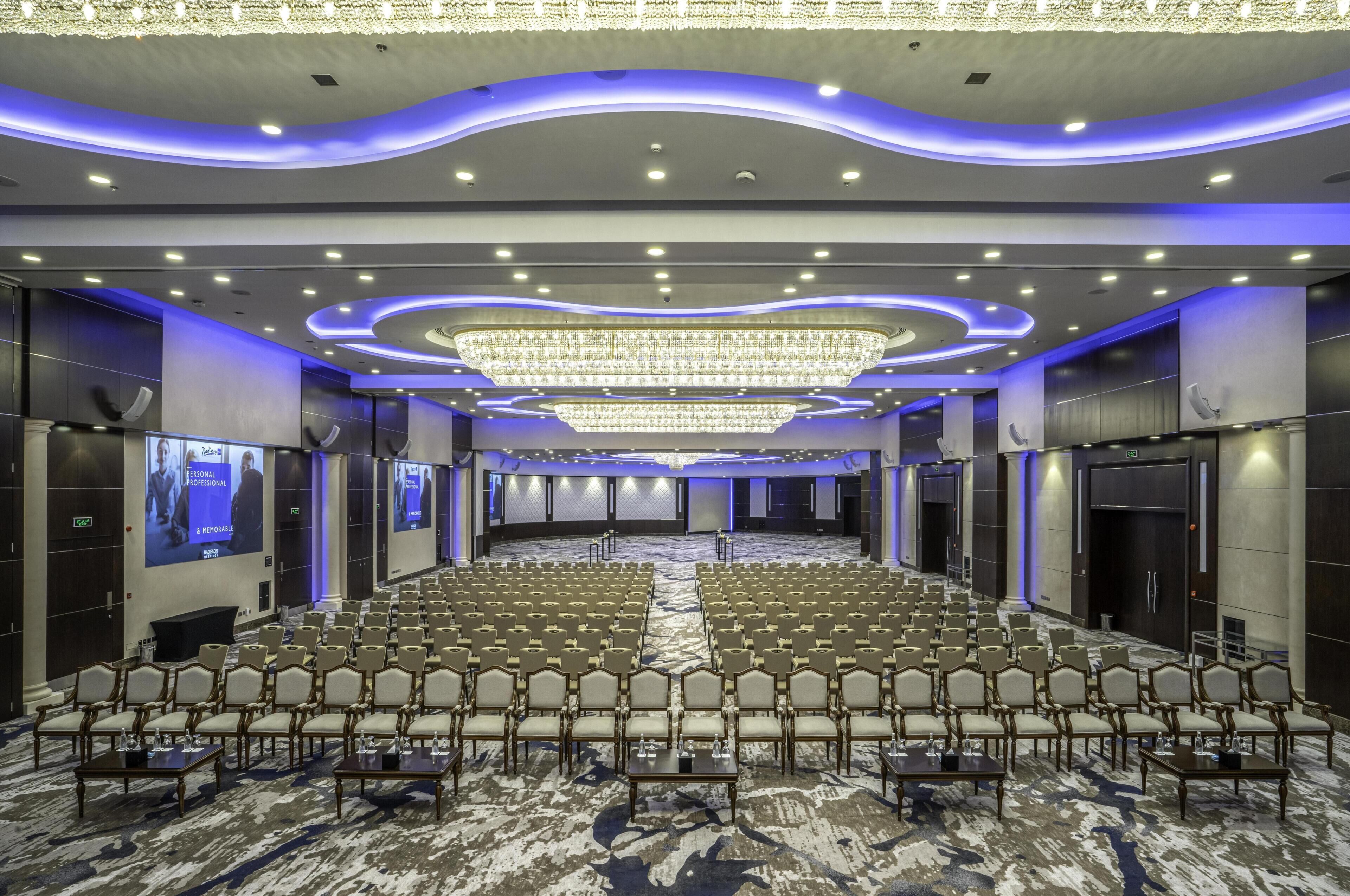 Een ruime congreszaal met rijen stoelen, een centraal podium en een kroonluchter.