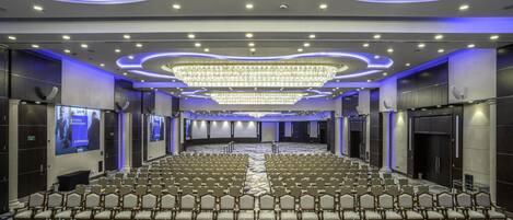 A large conference room with rows of chairs, a central stage, and a chandelier.