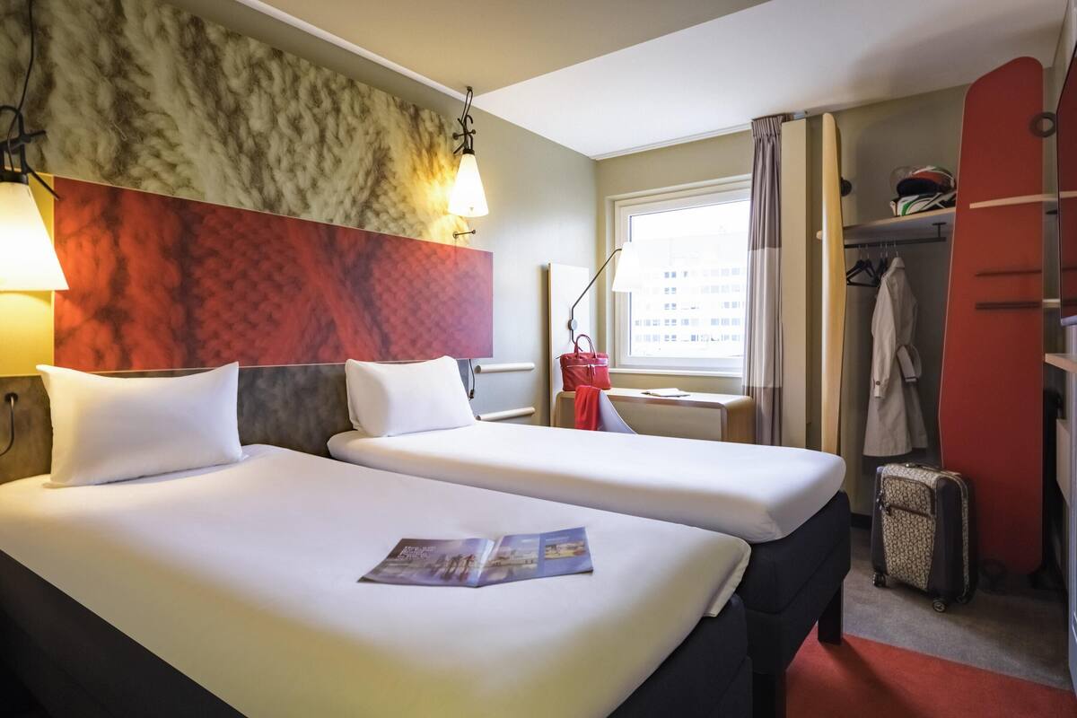 A hotel room with two beds, a red and white patterned headboard, a window with curtains, a coat rack, and a suitcase.