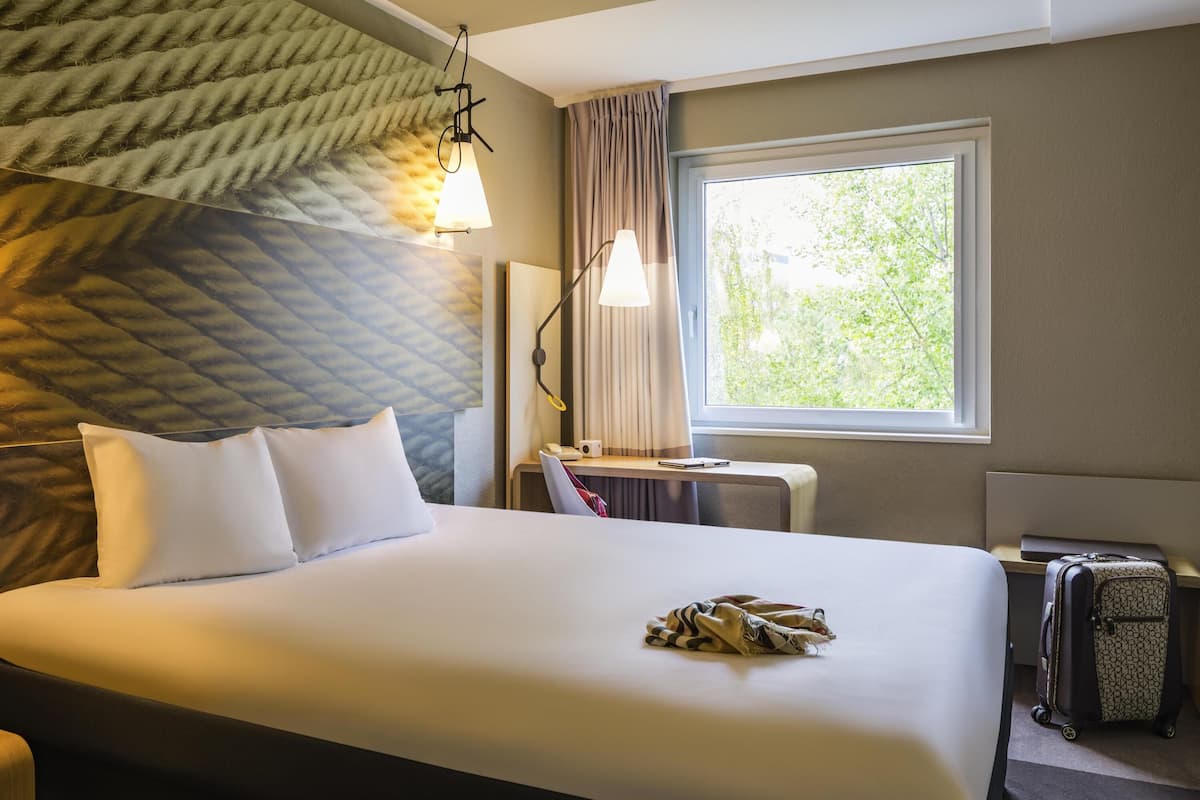 A hotel room with a bed, a desk, a lamp, a window, and a suitcase.