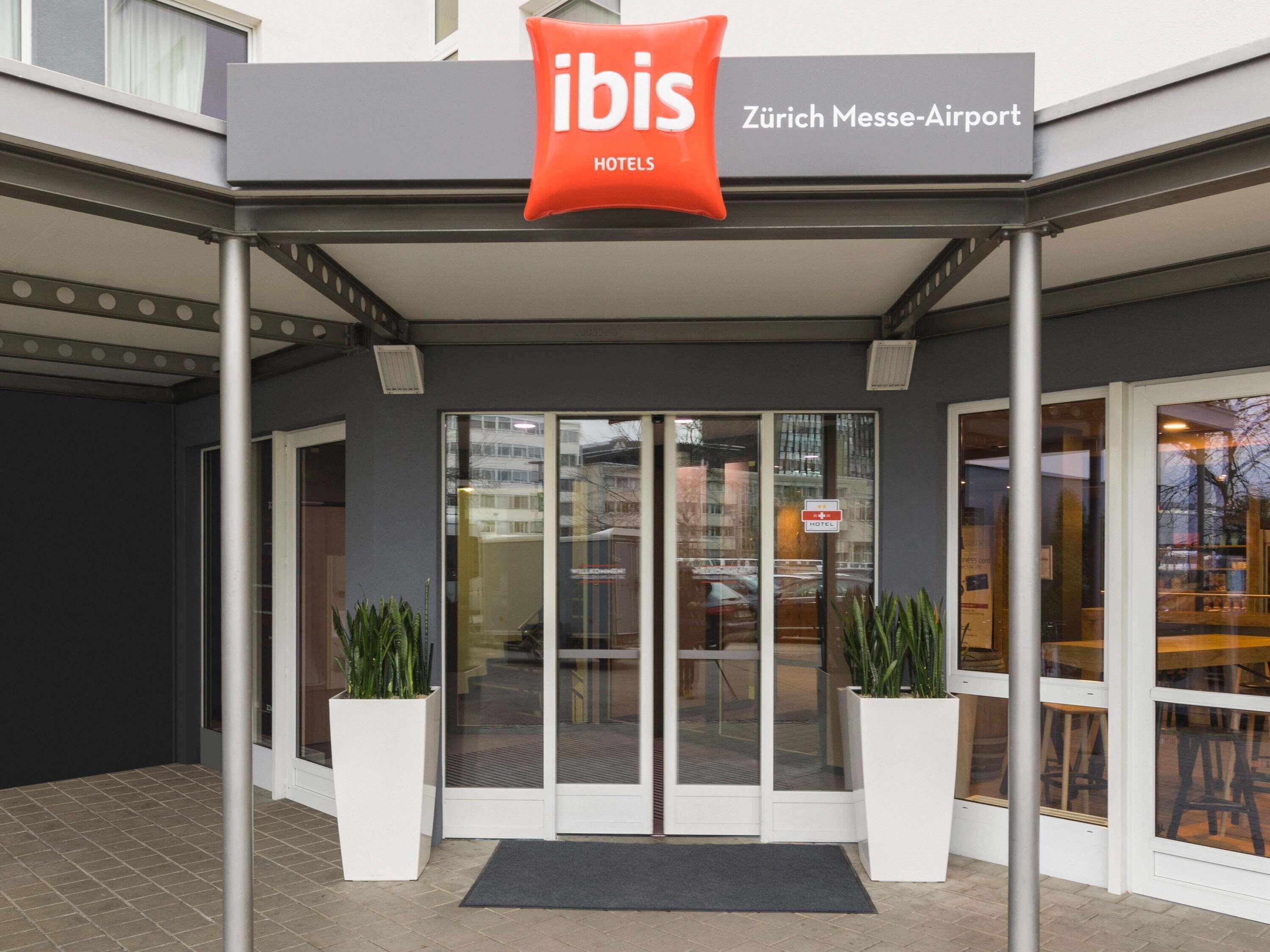 L’entrée des hôtels ibis se caractérise par une porte en verre, des plantes en pot et un panneau indiquant l’emplacement, à savoir Zürich Messe-Airport.