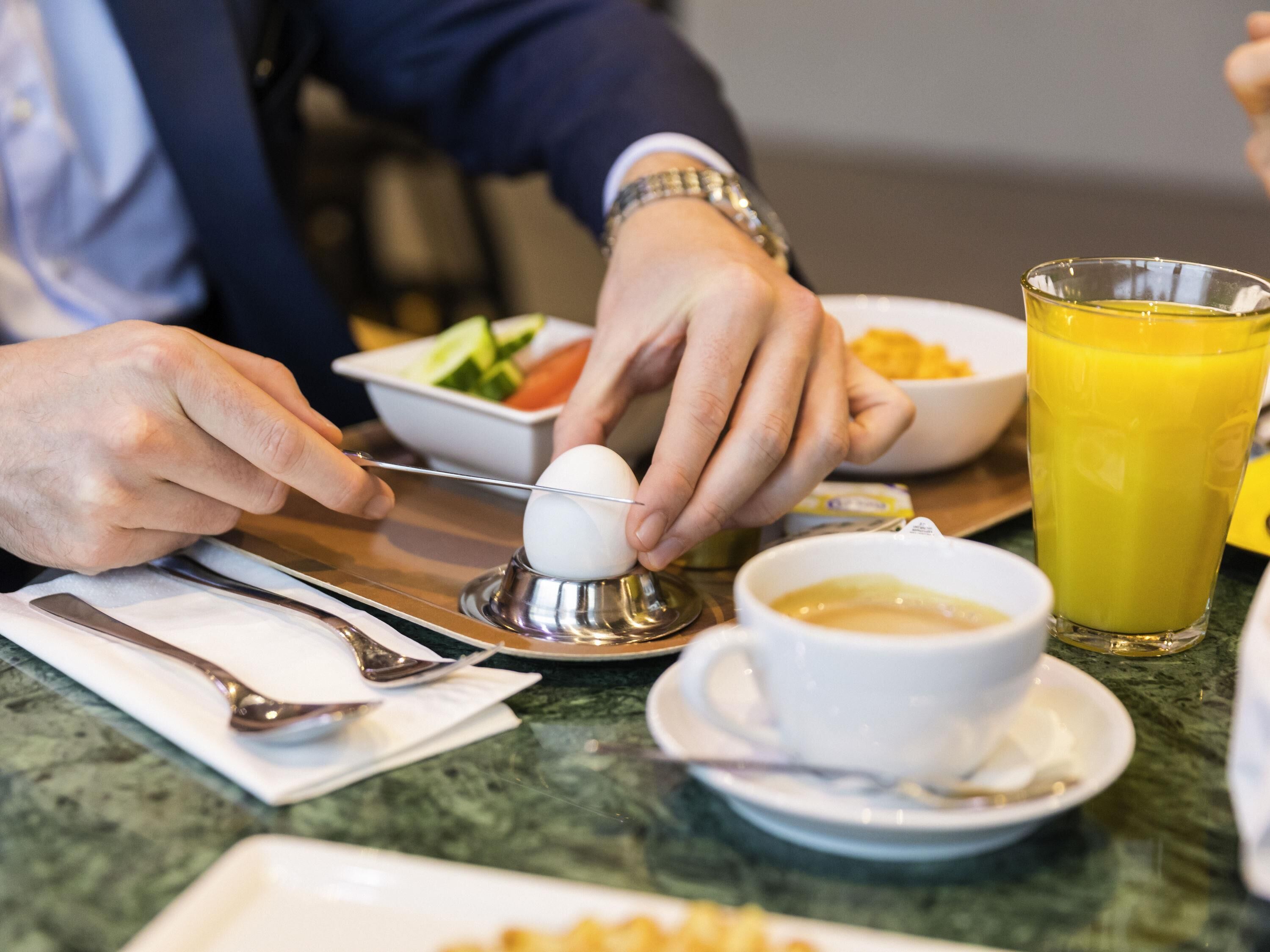 Un desayuno con una persona preparando un huevo, un tazón de fruta, un vaso de jugo de naranja y una taza de café sobre una mesa de mármol.