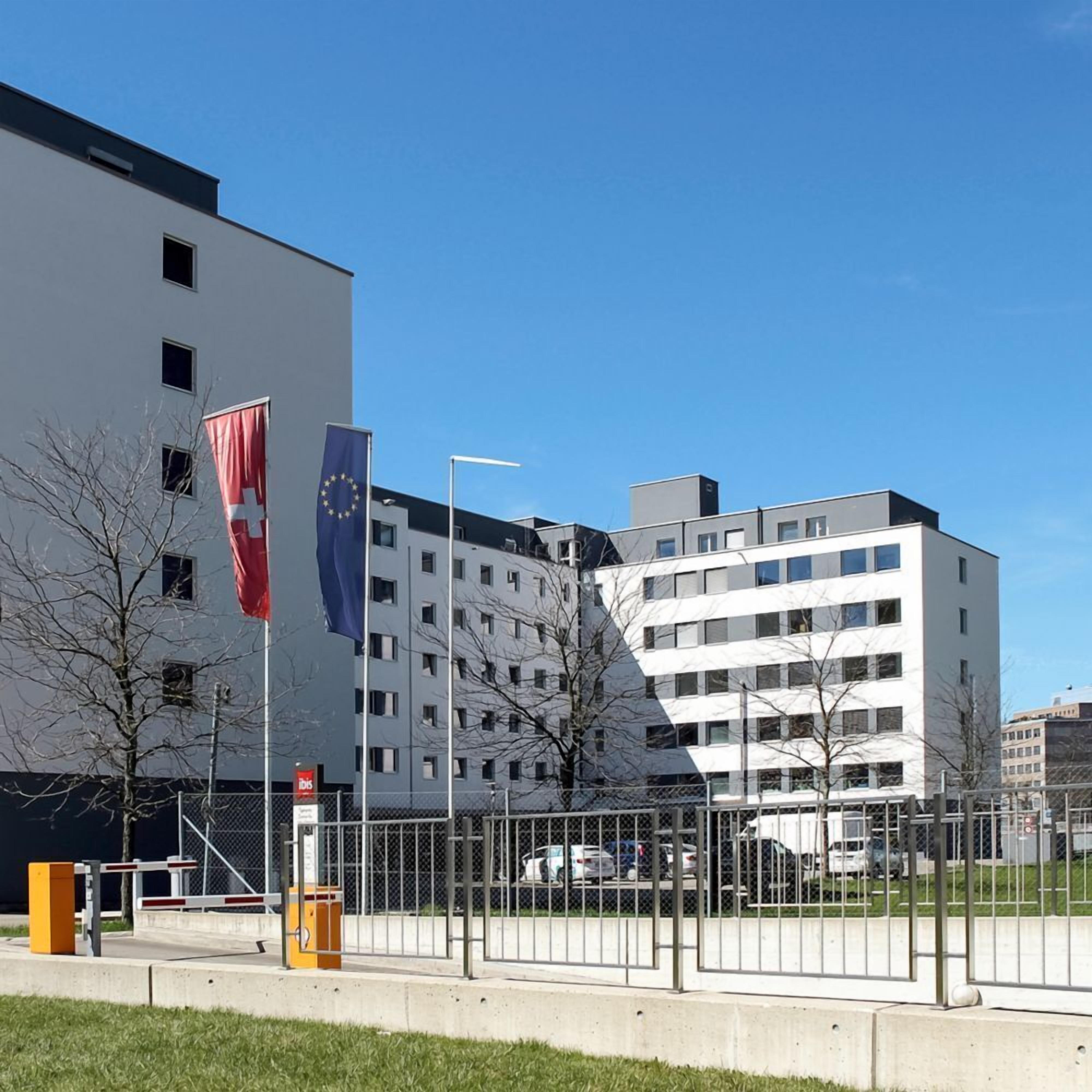 A modern building with multiple windows, a flagpole with a flag, and a fenced parking area.