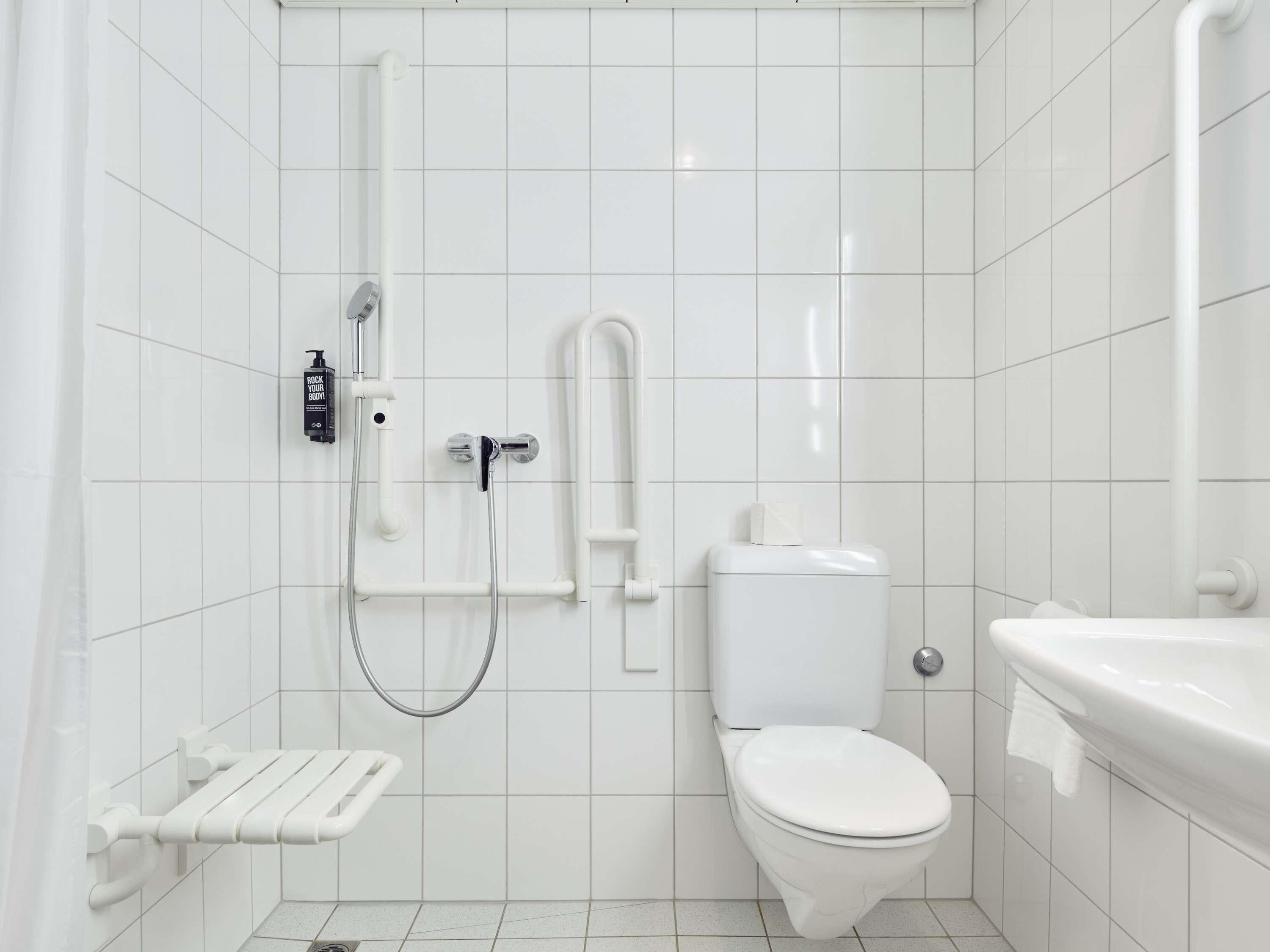 A small, modern bathroom with white tiles, a toilet, a sink, and a shower with a handheld showerhead.