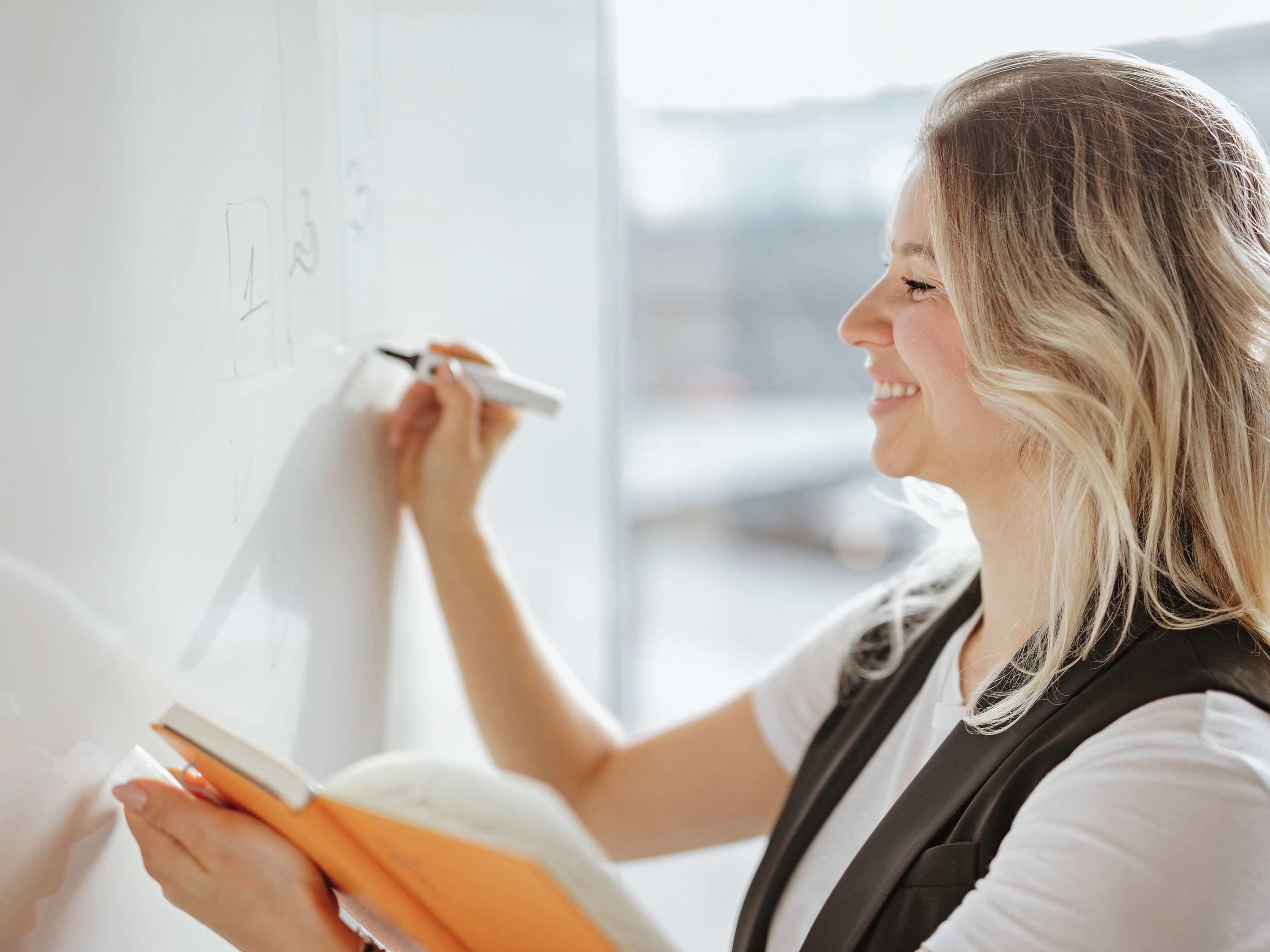 A person writing on a whiteboard with markers, holding a book.