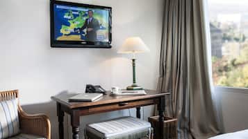 Camera d'albergo con televisione che mostra una previsione del tempo, una scrivania con lampada, un computer portatile e un telefono. Una finestra con tende offre vista all'esterno.