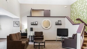 Una sala de estar moderna con un sofá, un sillón y una mesa de centro. Un televisor de pantalla plana está montado sobre un mueble bajo, y se ve una escalera al fondo.