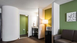 Habitación de hotel con kitchenette, sofá, lámpara y un cuadro en la pared.