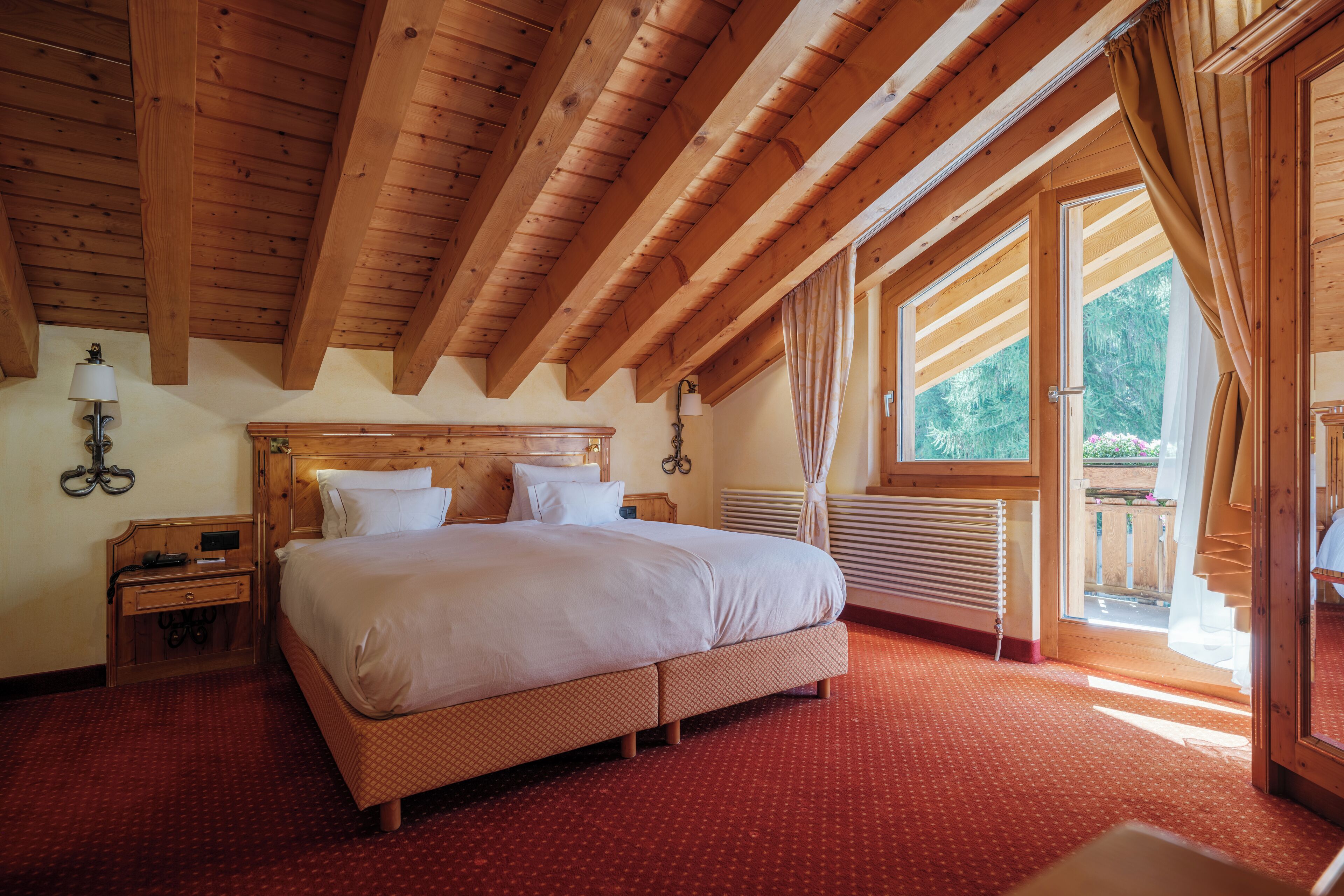 A wooden-framed bedroom with a large bed, a nightstand, a lamp, and a window with curtains.