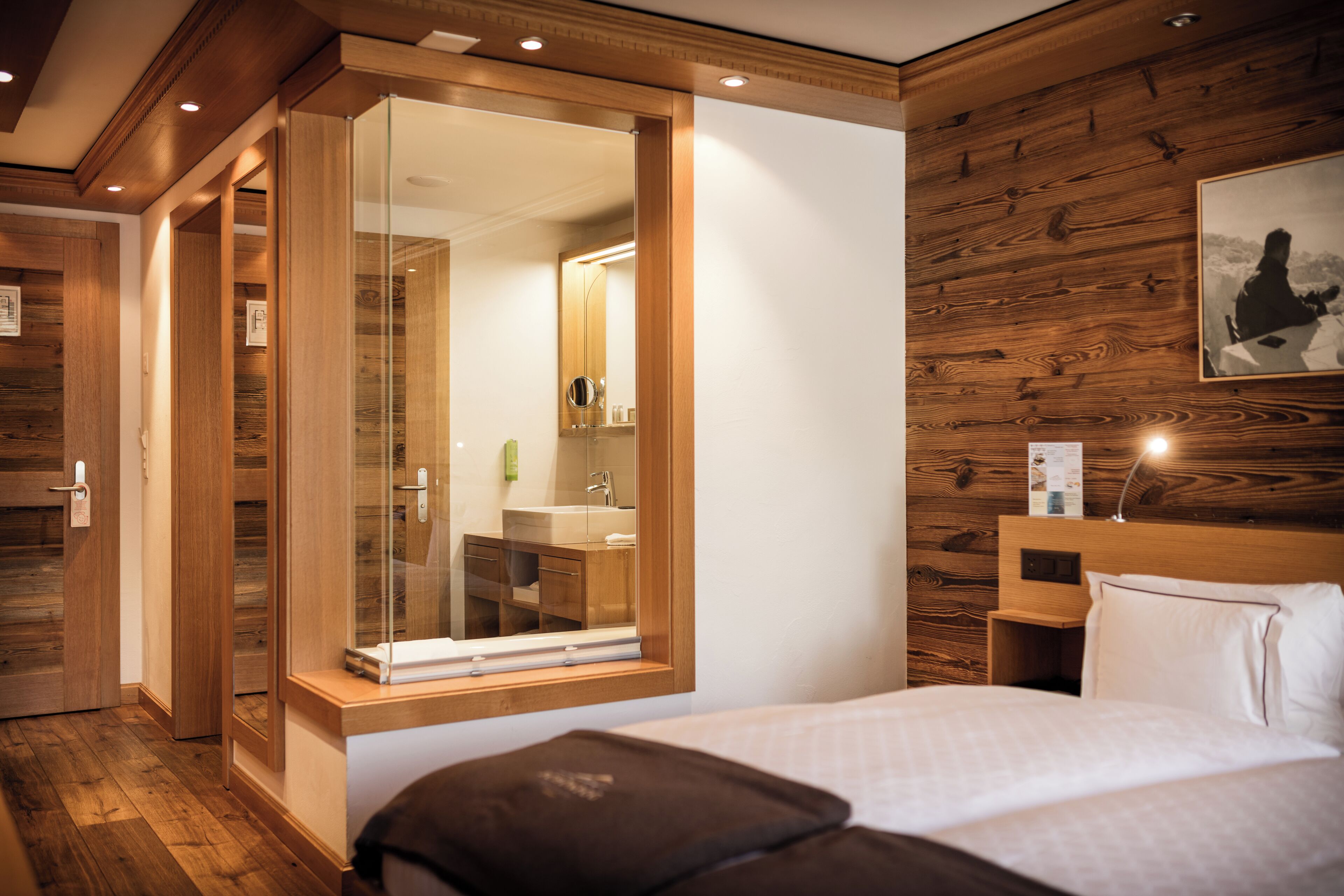Habitación de hotel con pared de madera, un espejo grande, una cama con ropa blanca y una mesita de noche de madera.