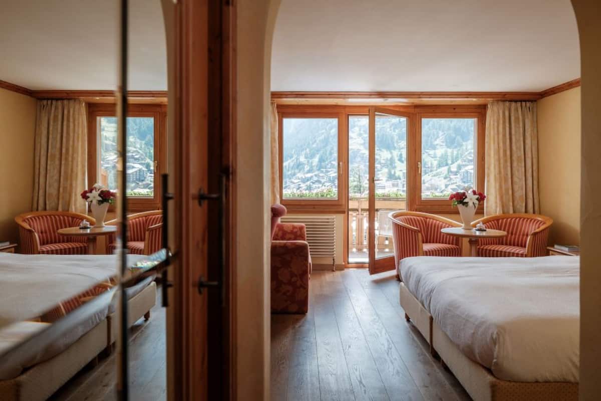 A hotel room with two beds, a sitting area with chairs, a small table, and a view of mountains through large windows.