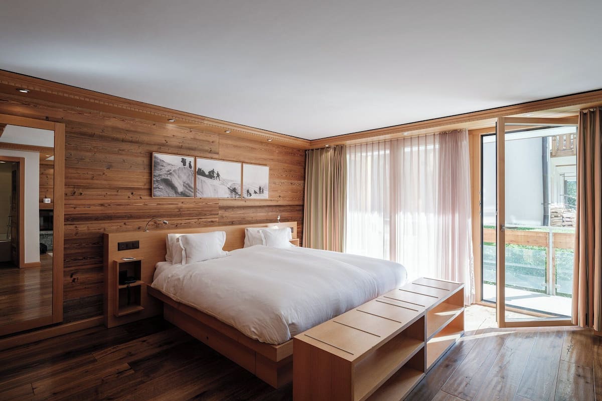 A modern bedroom with a wooden headboard, a large bed, a bench, and a sliding glass door to the outside.