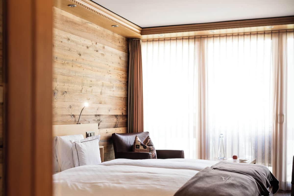A hotel room with a wooden headboard, a large bed with a brown blanket, a bedside table with a lamp, and a view of the outdoors through sheer curtains.