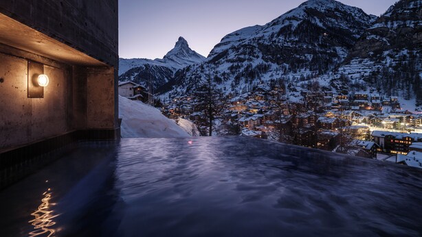 A snow-covered mountain town at dusk with a pool area featuring a stone wall and a light fixture. - Relais & Chateaux Schönegg (Zermatt)