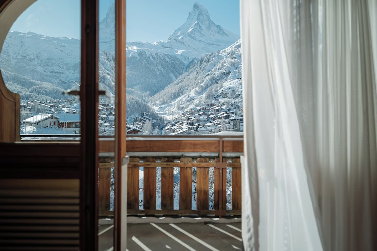 A view from a balcony with a wooden railing, overlooking a snow-covered mountain landscape with a prominent peak and a village below.