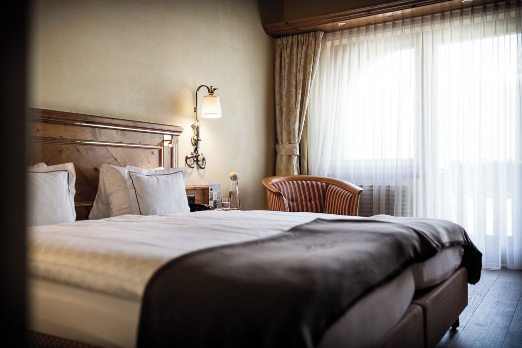 A hotel room with a large bed, a wooden headboard, a bedside table with a lamp, a chair, and a window with sheer curtains.