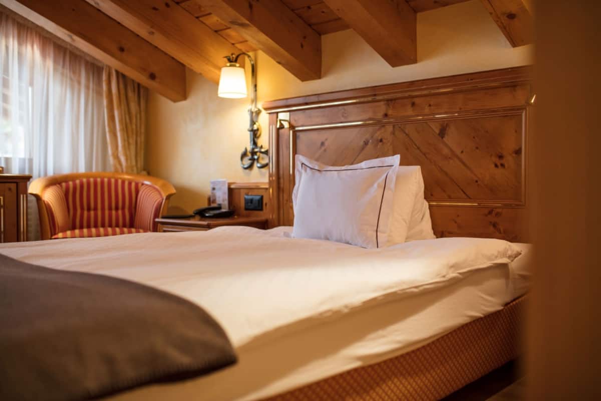 A hotel room with a wooden headboard, a bed with white linens, a striped armchair, and a nightstand with a lamp.