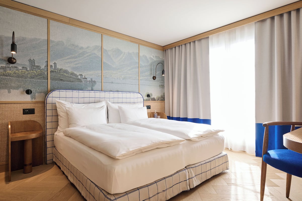 A hotel room with a large bed, a wooden nightstand, a chair, and a view of mountains through a window.