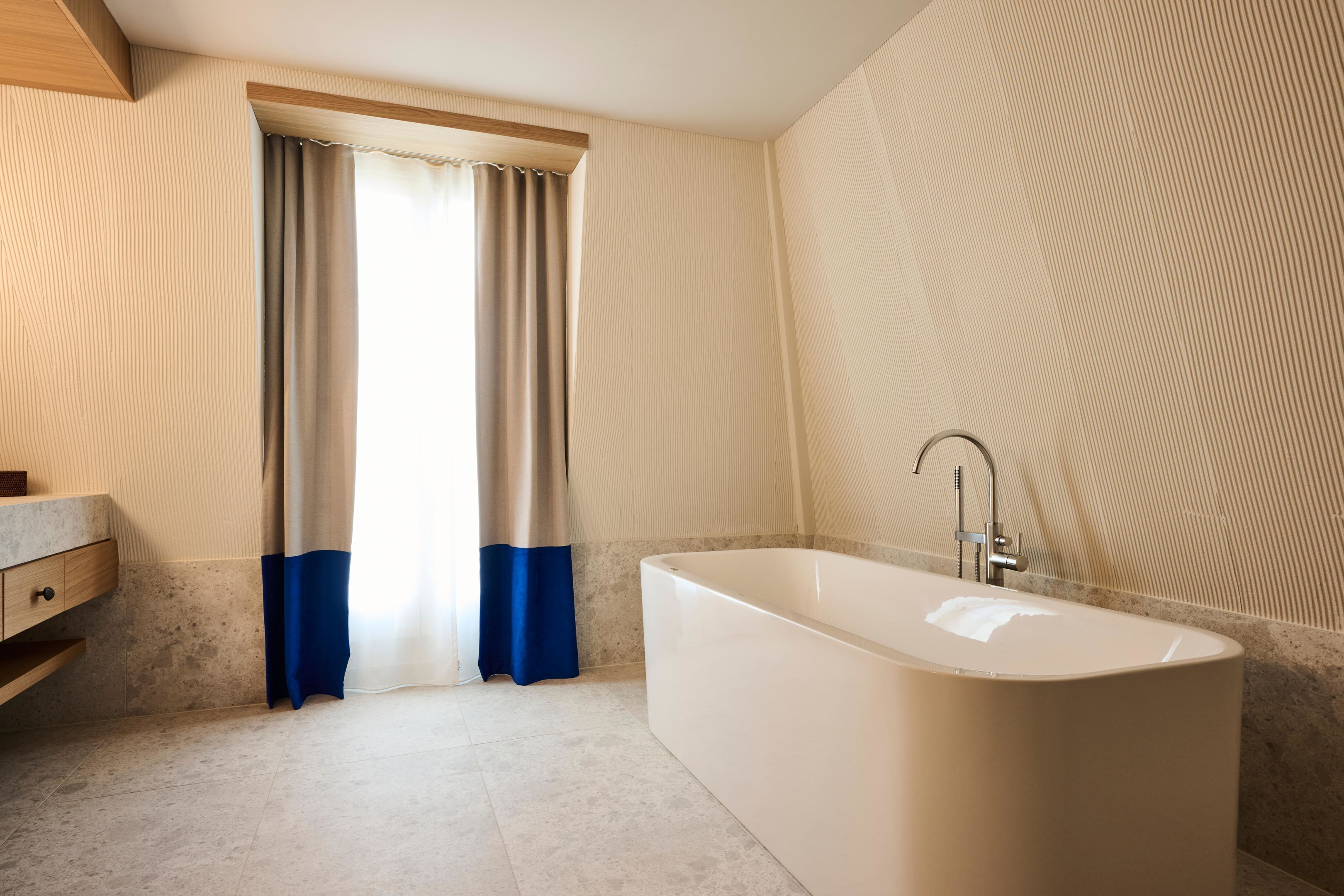 Een moderne badkamer met een vrijstaand bad, een wastafel met een marmeren blad en een groot raam met gordijnen.