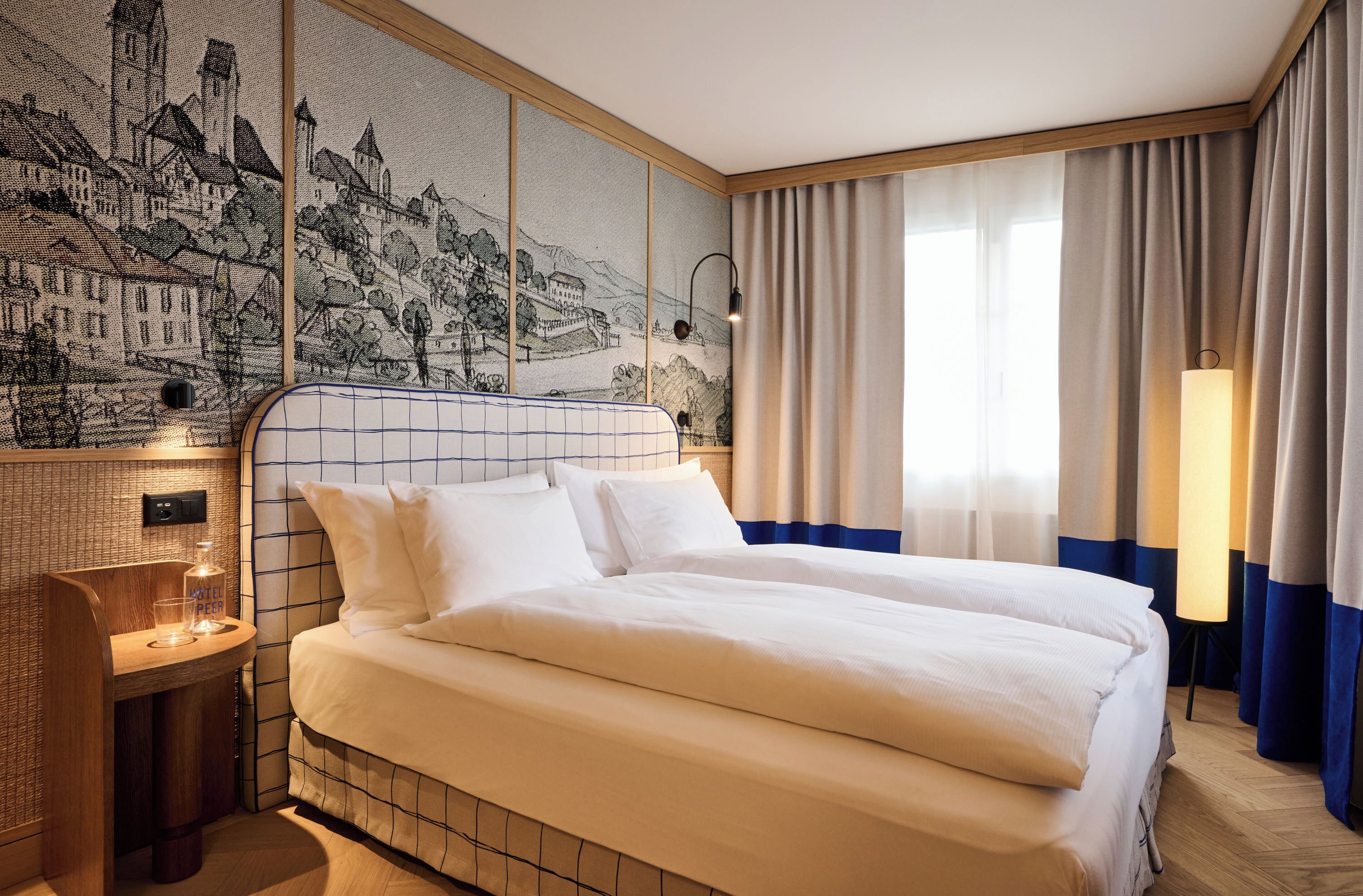 A hotel room with a large bed, a bedside table with a bottle and glass, a wall tapestry, and a window with curtains.