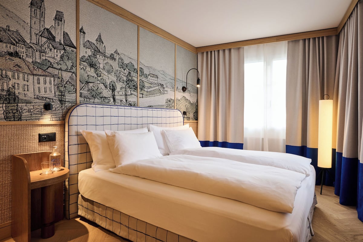 A hotel room with a large bed, a small bedside table with a bottle and glass, a wall-mounted light, and a large window with curtains.
