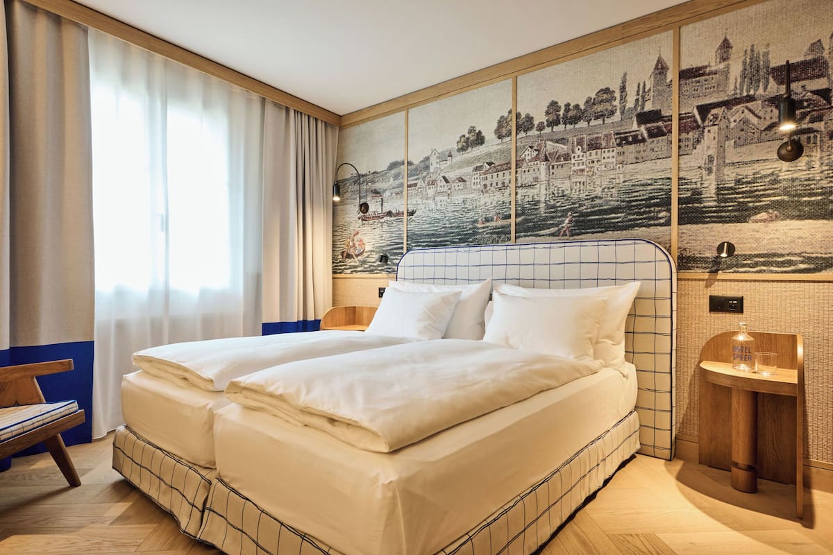A hotel room with a large bed, a wooden headboard, a nightstand with a lamp, and a view of a river scene on the wall.